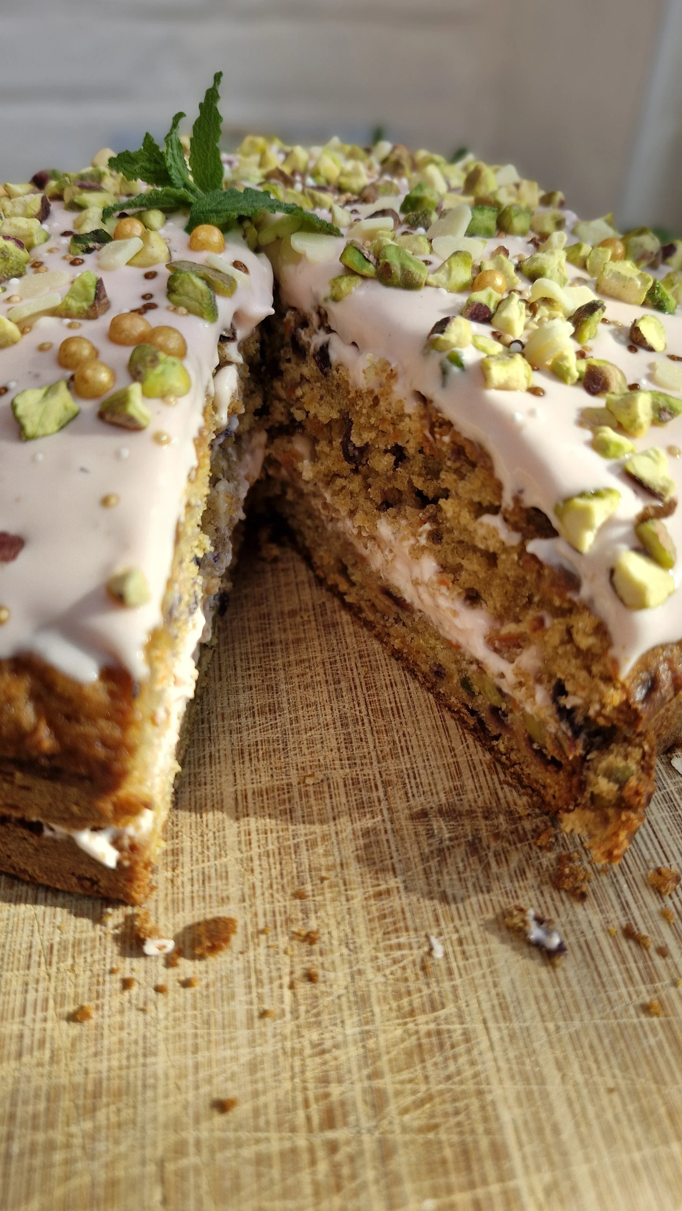 Close-up van een stuk taart met roomkaas icing, versierd met gehakte pistachenoten en een muntblaadje, op een houten snijplank.