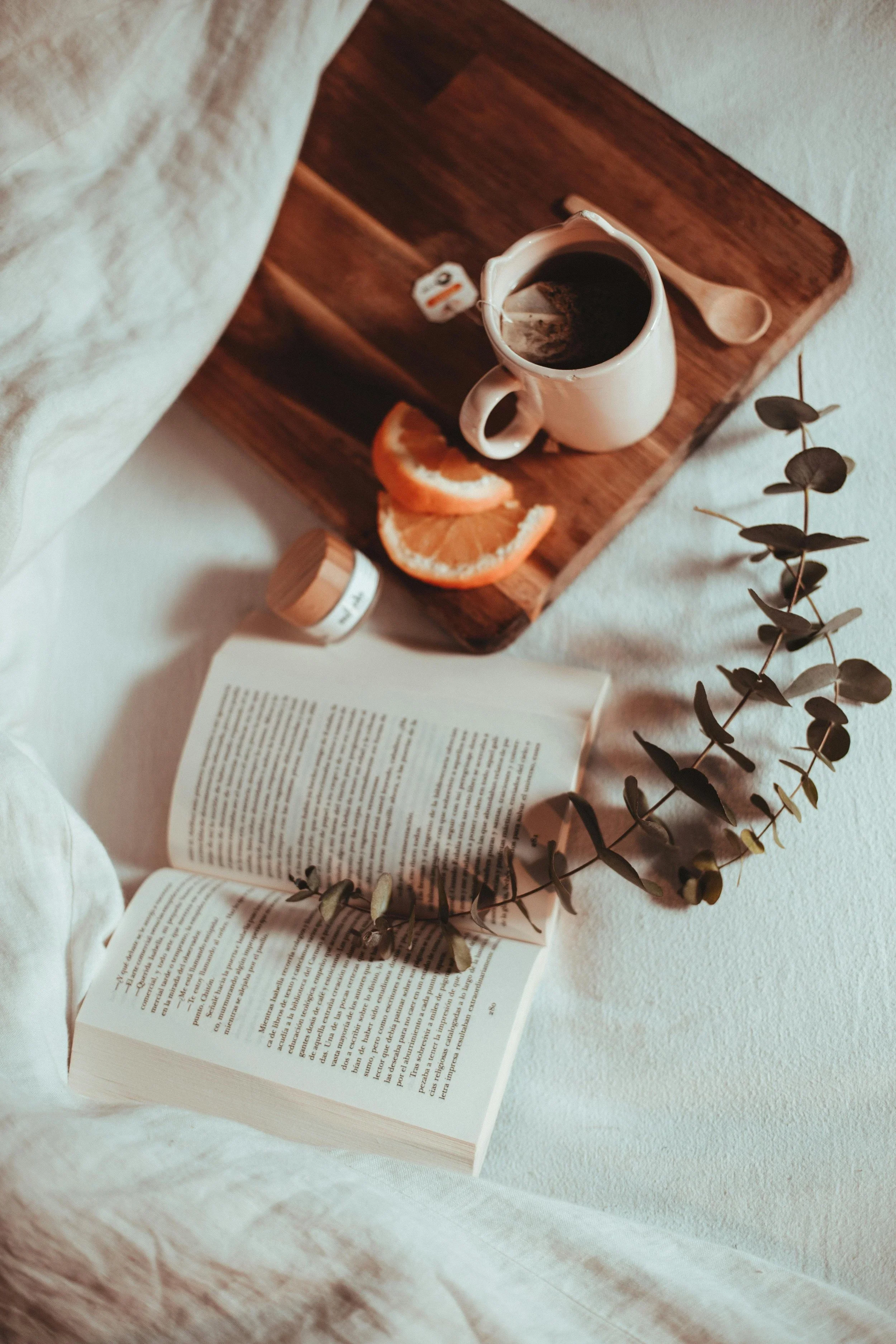 Een open boek ligt op een zachte, crèmekleurige stof. Daarnaast ligt een houten snijplank met een kopje zwarte koffie, een schijfje sinaasappel en een schatting van een theezakje. Een groene plant met ronde bladeren ligt langs de rand van de afbeelding.
