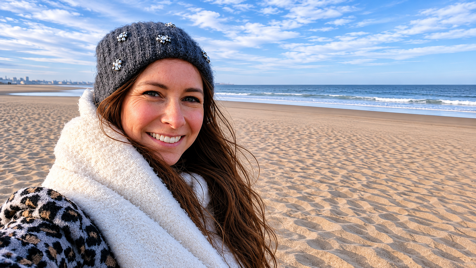 Een glimlachende vrouw met lang bruin haar, gekleed in een dikke witte sjaal en een grijze muts met bloemen, staat op een zandstrand bij de oceaan onder een blauwe lucht met wolken.