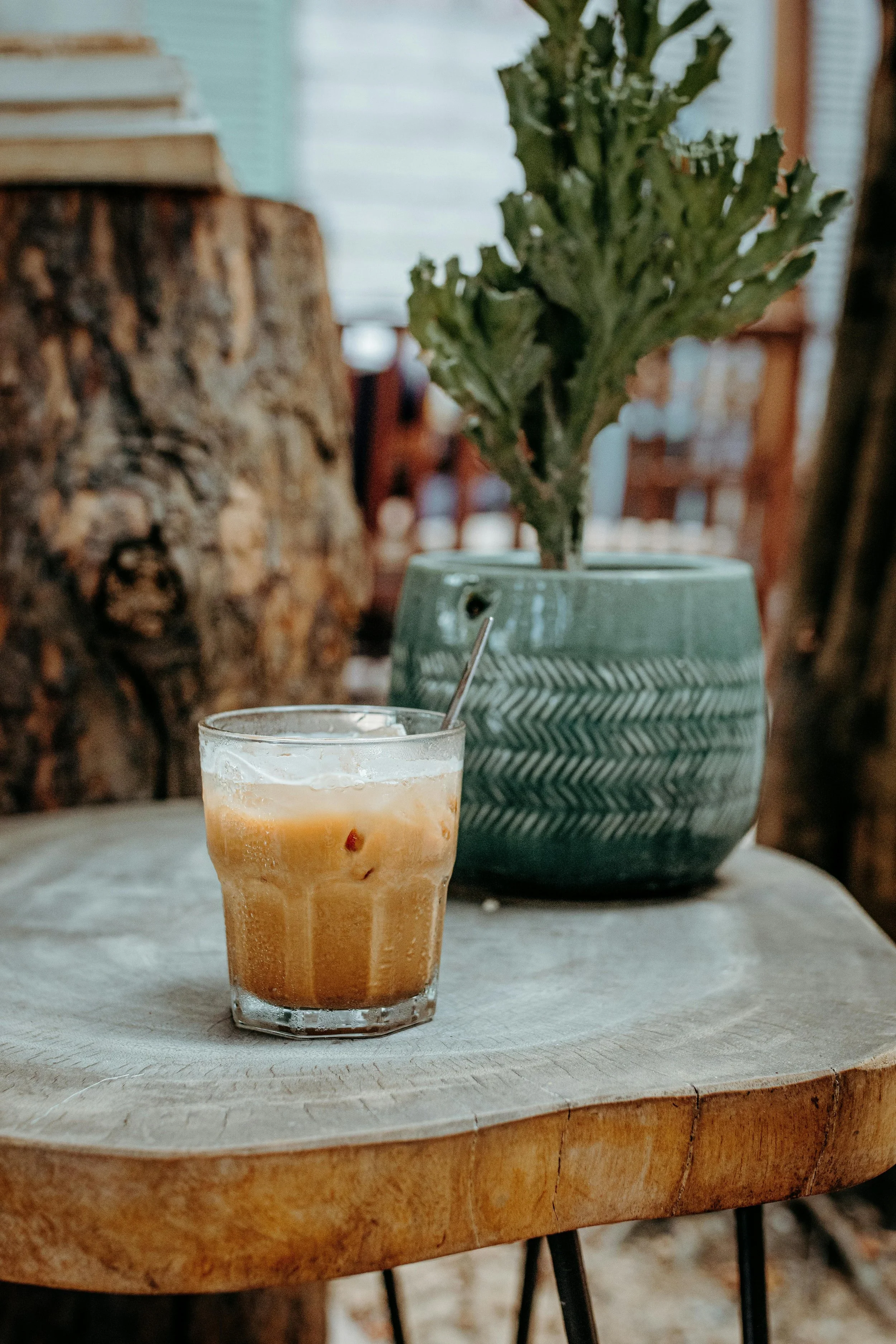 Een glas met een ijsdrankje op een houten tafel, op de achtergrond een groene vaas met een plant, en een plank met hout. Ruimte voor verdere context.