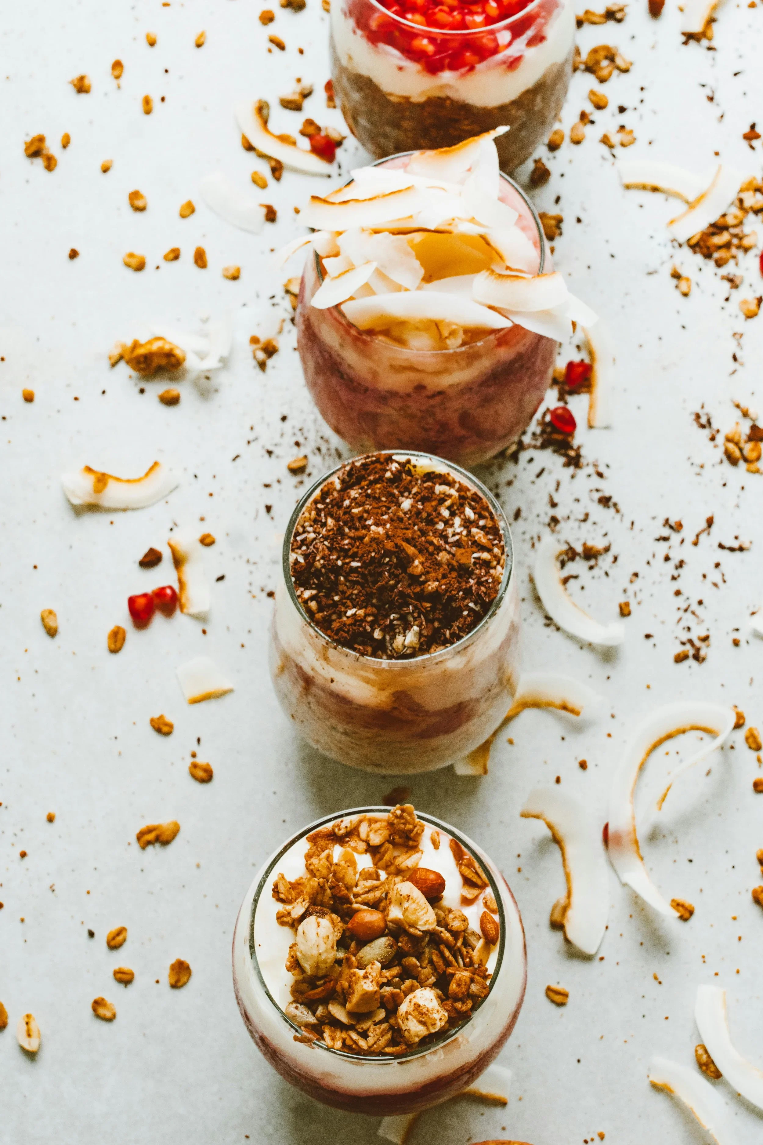 Drie glazen met verschillende soorten nootjes en granola, omringd door scattered nootjes, granola en kokosvlokken op een witte achtergrond.