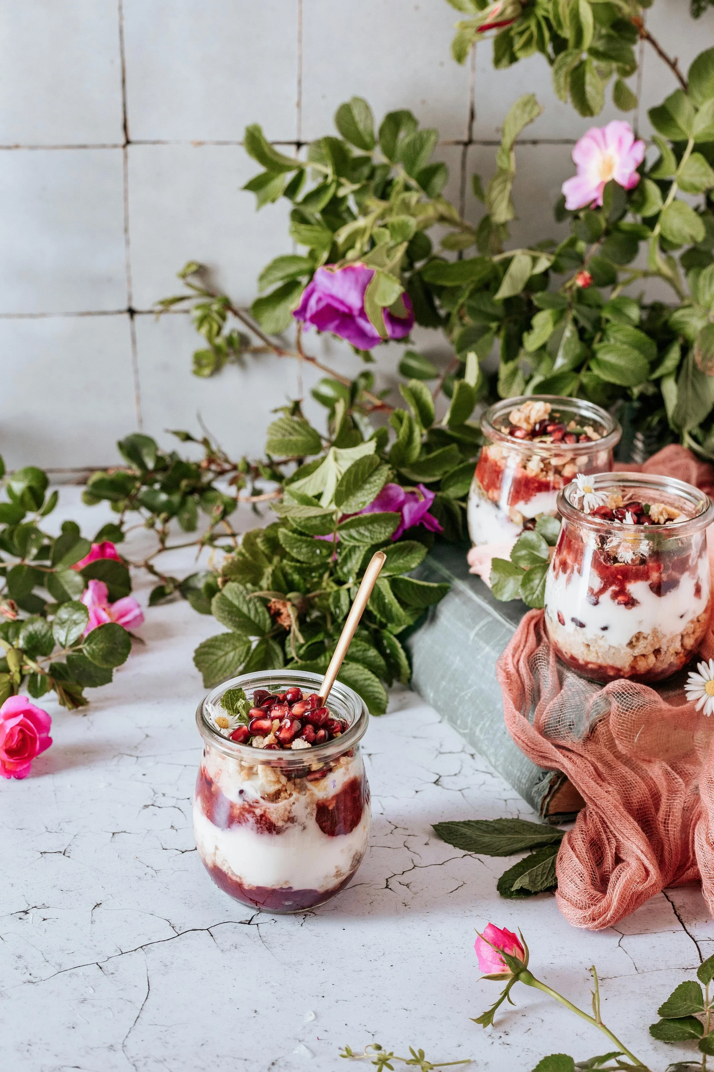 Glas met yoghurt, rood fruit, granola en bloemen, omgeven door groene bladeren en roze bloemen.