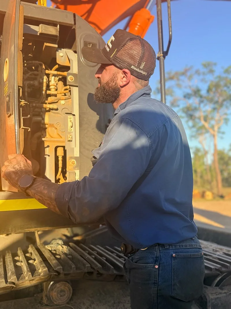 Owner of CMS Jake wearing a cap, long-sleeve shirt, and a watch, working on the engine of heavy construction equipment outdoors during daytime.