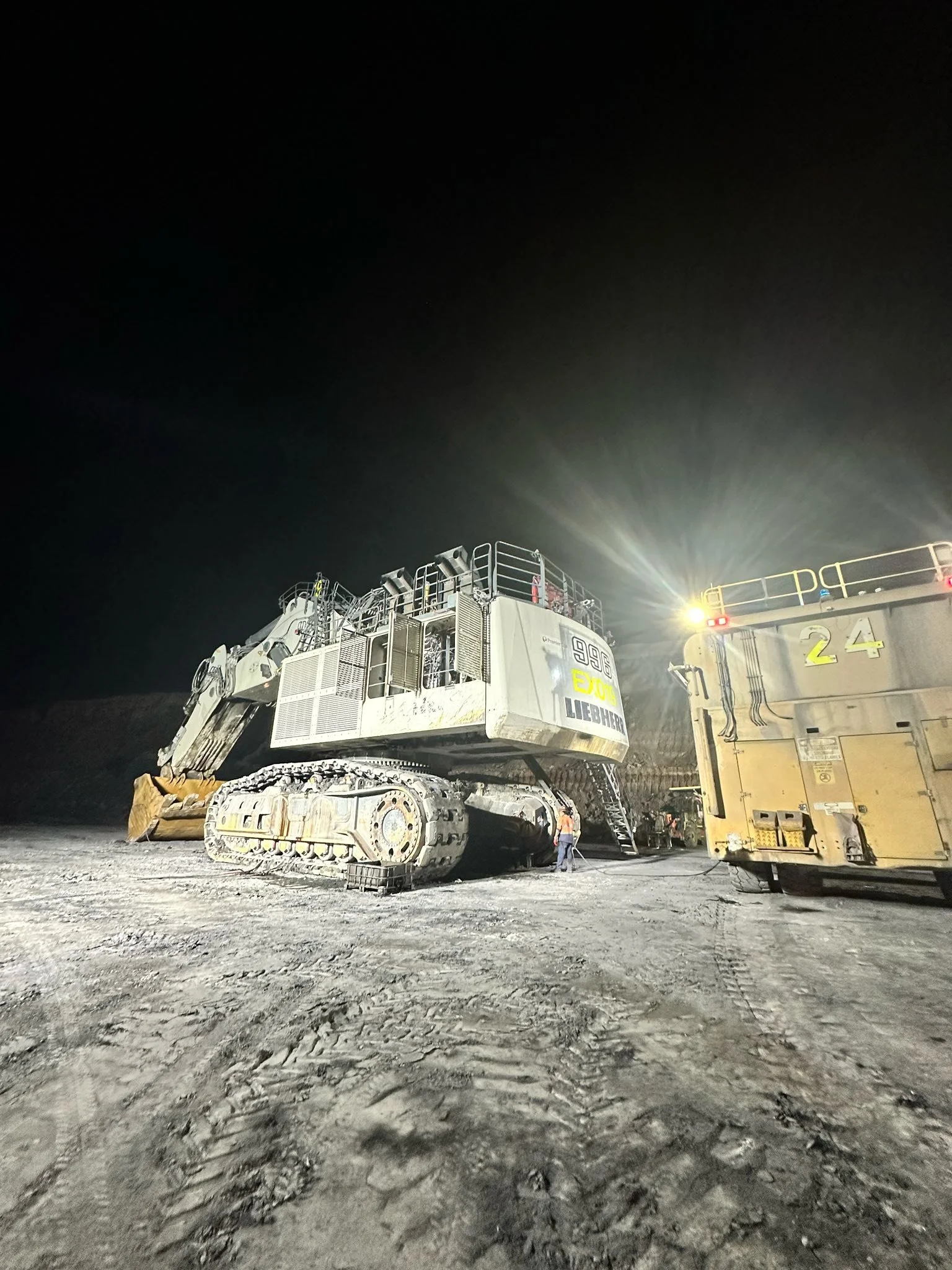 Nighttime photo of a large construction vehicle, possibly a bulldozer, on a dirt surface with tire tracks, illuminated by bright artificial lights.