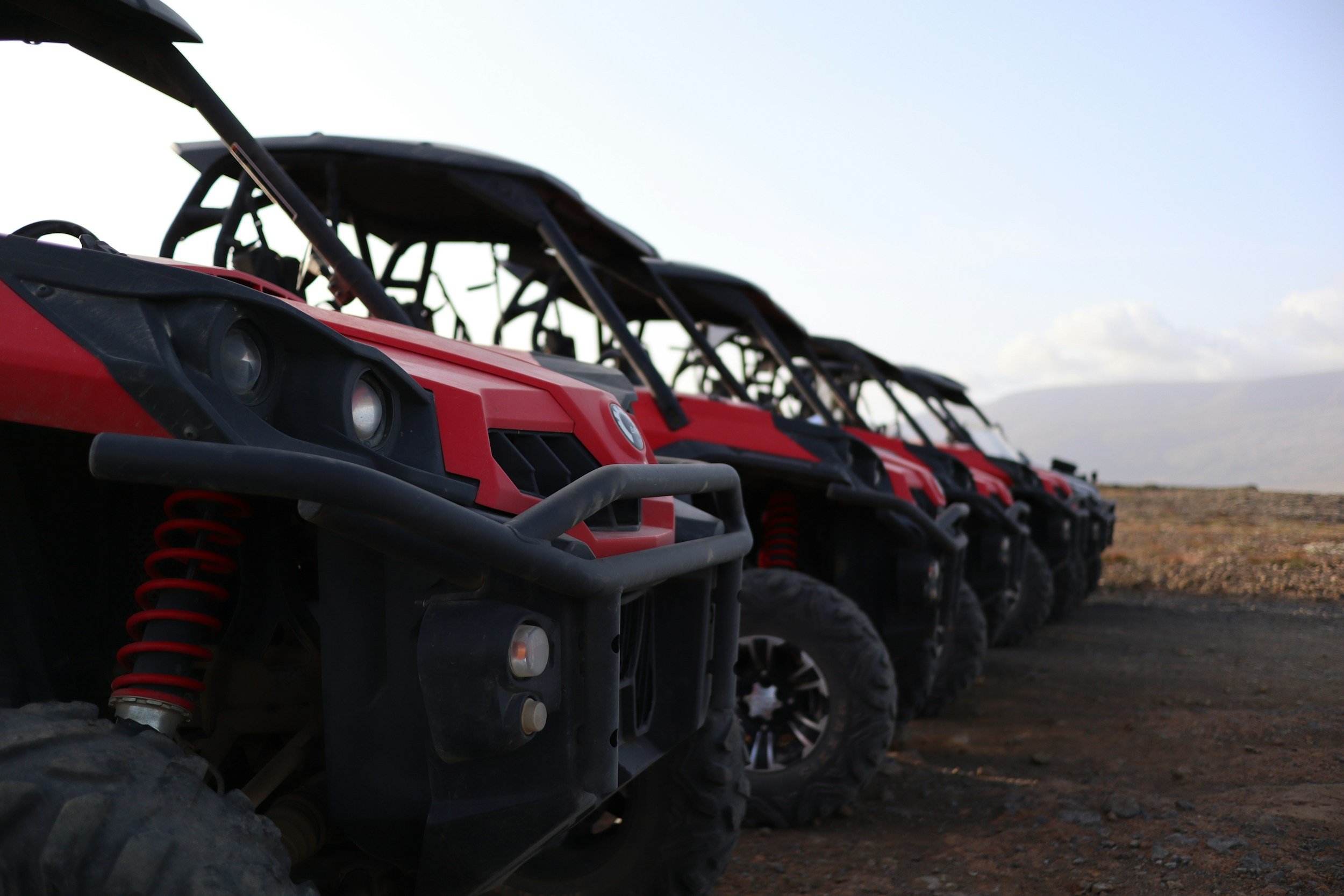 A row of red off-road vehicles with black roll cages, large tires, and suspension systems parked on a dirt surface outdoors under a cloudy sky.