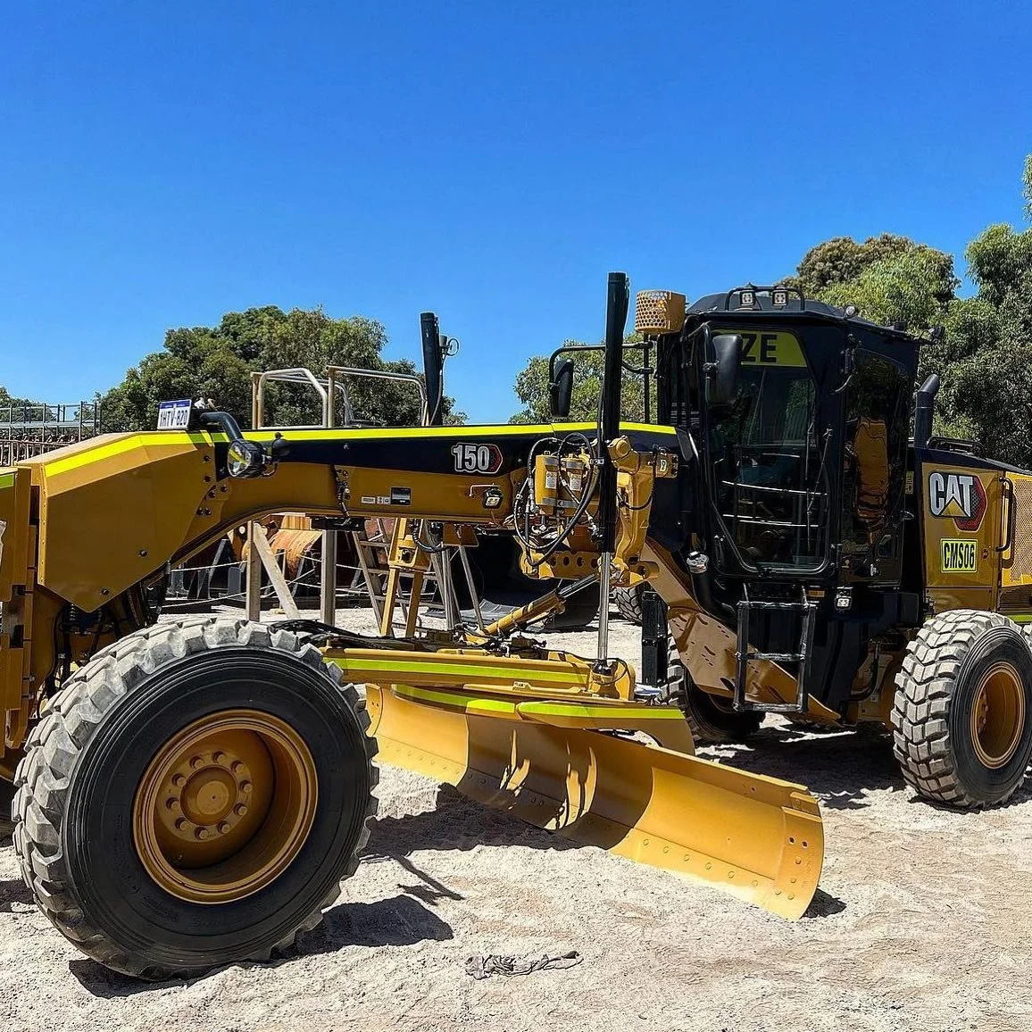 Yellow and black Caterpillar road grader on construction site with blue sky and trees in the background.