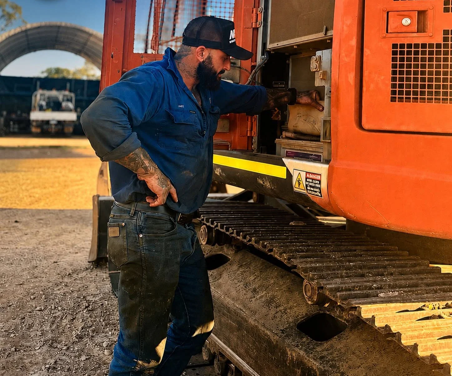 Man in blue work clothes with tattoos on his arm, wearing a black cap, working on an orange excavator.