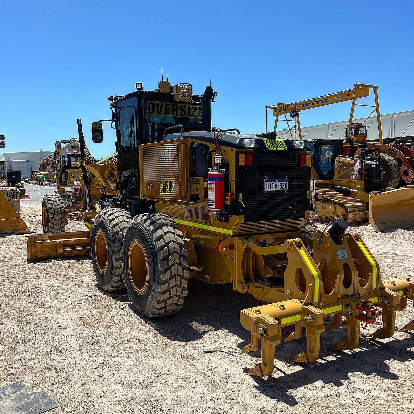 Yellow construction vehicle with large tires and boom attachment, parked on dirt lot under clear blue sky, with other heavy equipment in background.