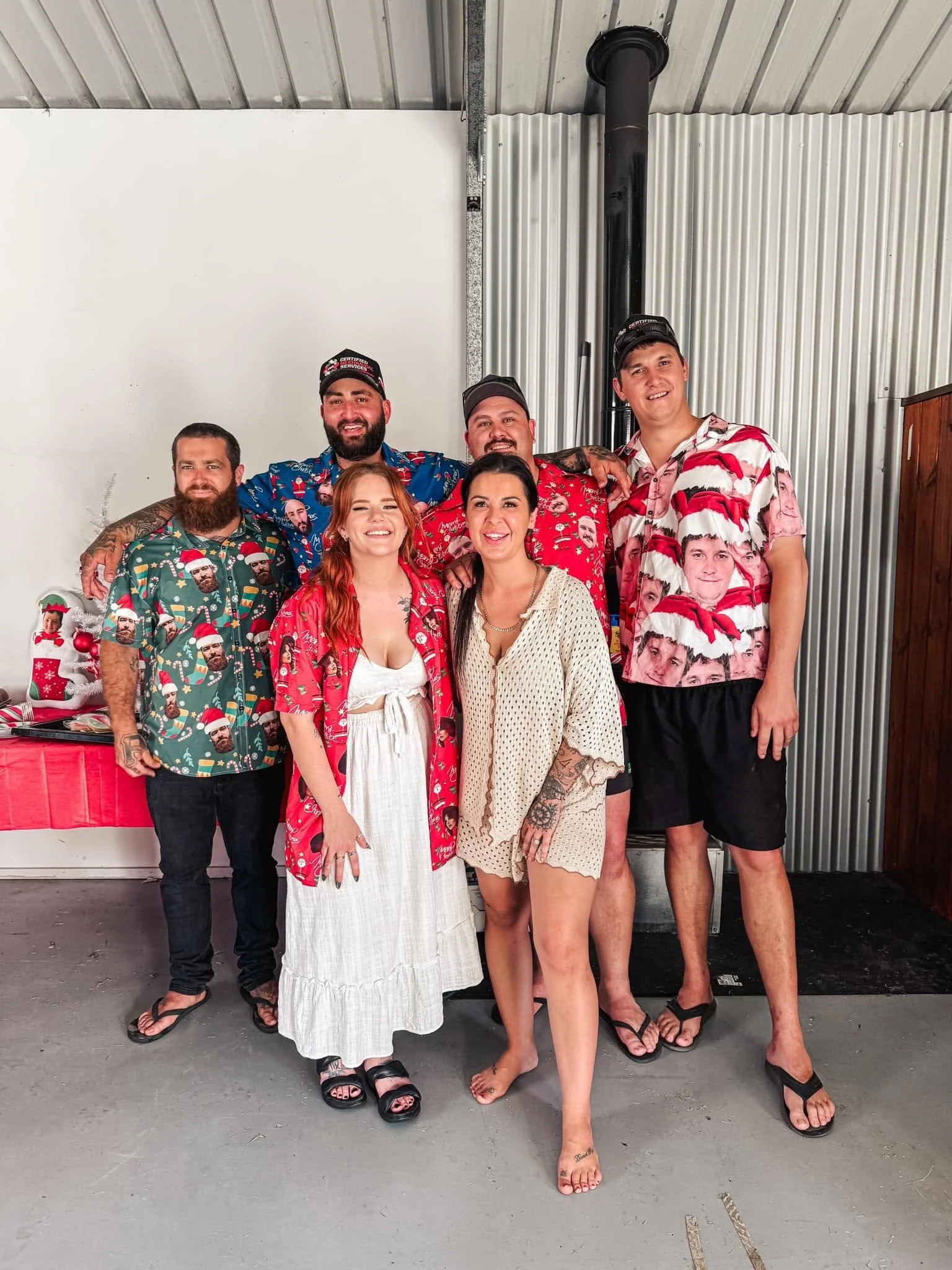 A group of seven people standing together indoors, celebrating Christmas, wearing festive shirts with faces and holiday themes, some smiling, some with relaxed expressions, against a background with a white wall, corrugated metal, and a black chimney pipe.