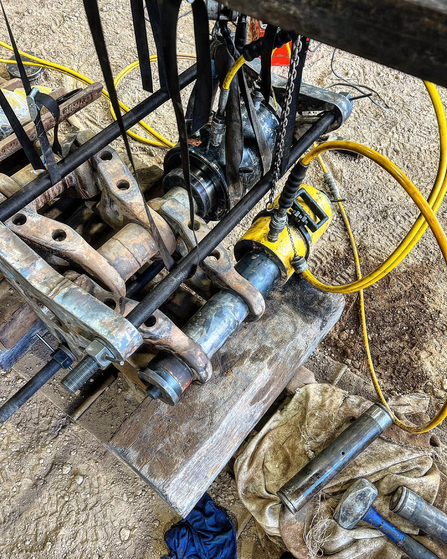 Close-up of a construction or industrial machinery with exposed metal parts, hoses, wires, and a yellow tool, resting on a weathered wooden surface with dirt and debris around.