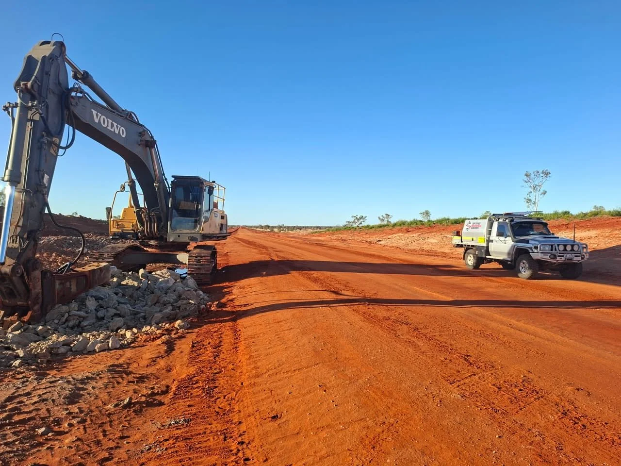 Construction site with a large excavator and a pickup truck on a dirt road under a clear blue sky.