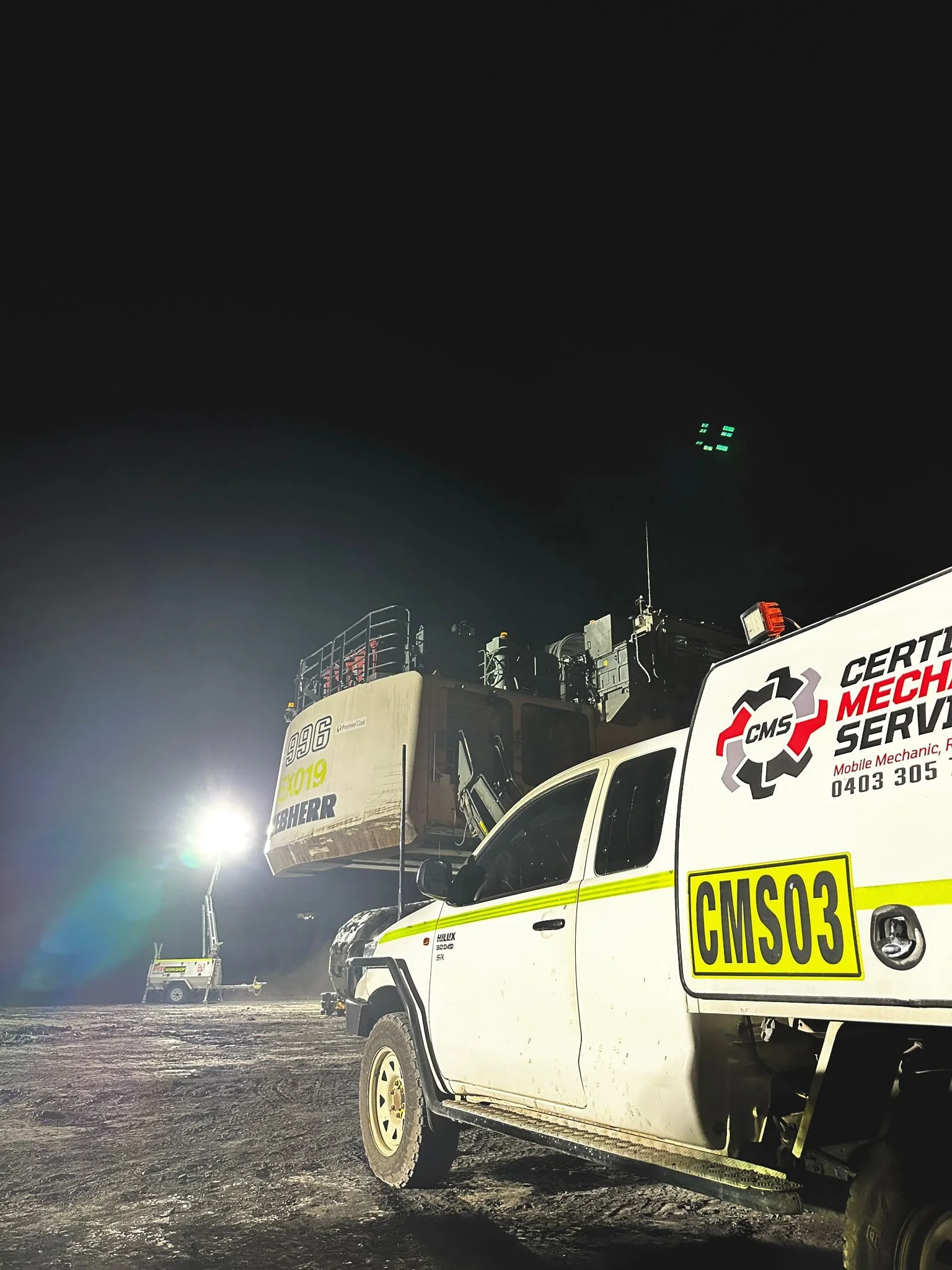 At night, a white utility truck with yellow reflective markings, labeled 'CMS03', is parked on a muddy surface. Behind it, a radar or communication device is mounted on a truck outfitted for technical services. In the background, there are additional trucks and a tall bright light illuminating the area.