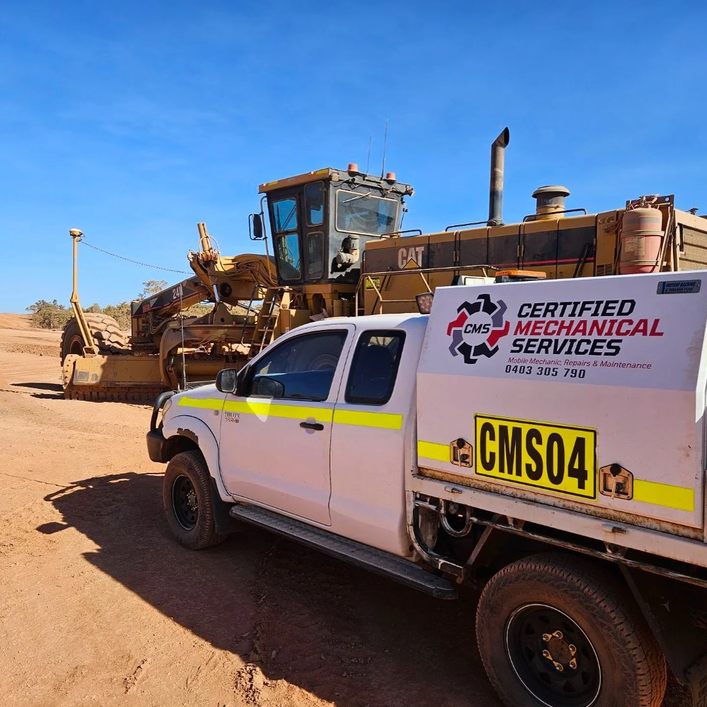 A white service truck with yellow stripes and the license plate CMS04 parked on a dirt surface next to large construction equipment, with a clear blue sky in the background.