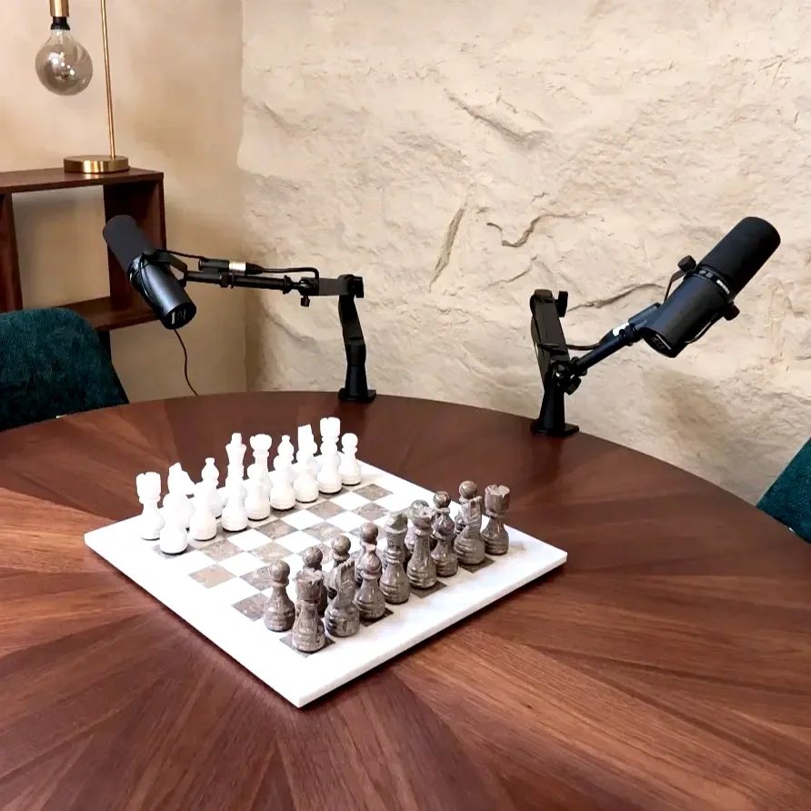 Set at Studio Analogue, Singapore Boutique Podcast studio with A chessboard with white and black chess pieces set up for a game on a wooden table. Two microphones are positioned above the table, and a table lamp is on a shelf in the background.
