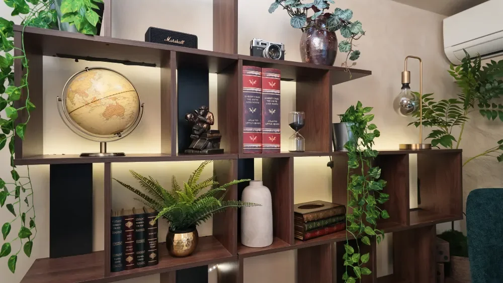 Interior room with wooden bookshelf decorated with plants, books, a globe, an hourglass, a sculpture, an air freshener, a camera, and a lamp.