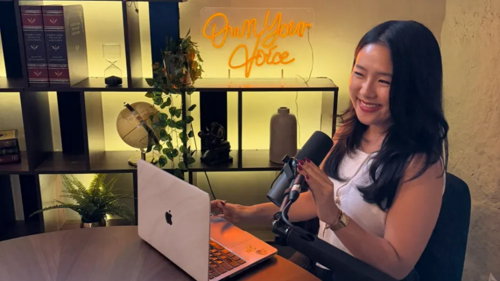 A woman with long dark hair smiling and holding a microphone while sitting at a desk with a laptop, in a room decorated with plants and shelves. There is a neon sign that says 'Own Your Voice' in the background.