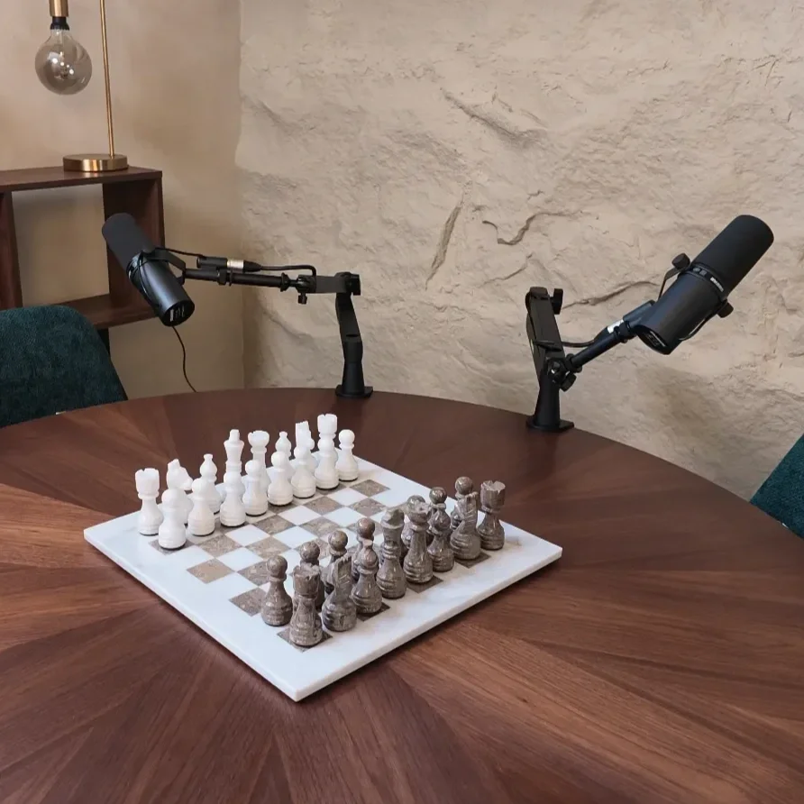 A chessboard with white and black chess pieces set up for a game, on a round wooden table at Studio Analogue, Singapore Boutique Podcast and vodcast recording studio. Two microphones on adjustable arms are positioned over the chessboard.