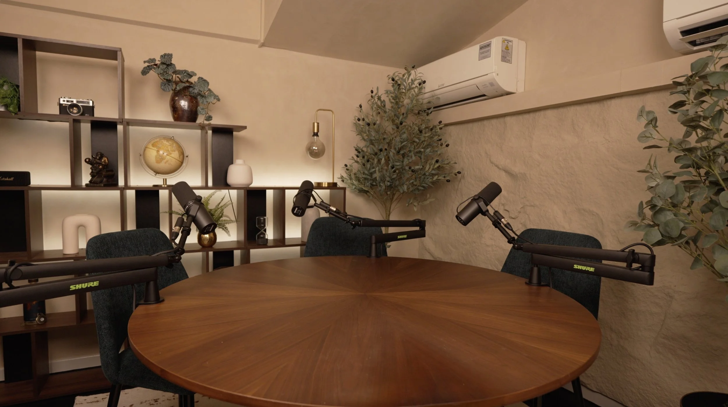 Studio Analogue, Singapore podcast recording studio with a round wooden table, three microphones, and fabric chairs. The background features a bookshelf with decorative objects and plants, a wall-mounted air conditioner, and a textured beige wall.