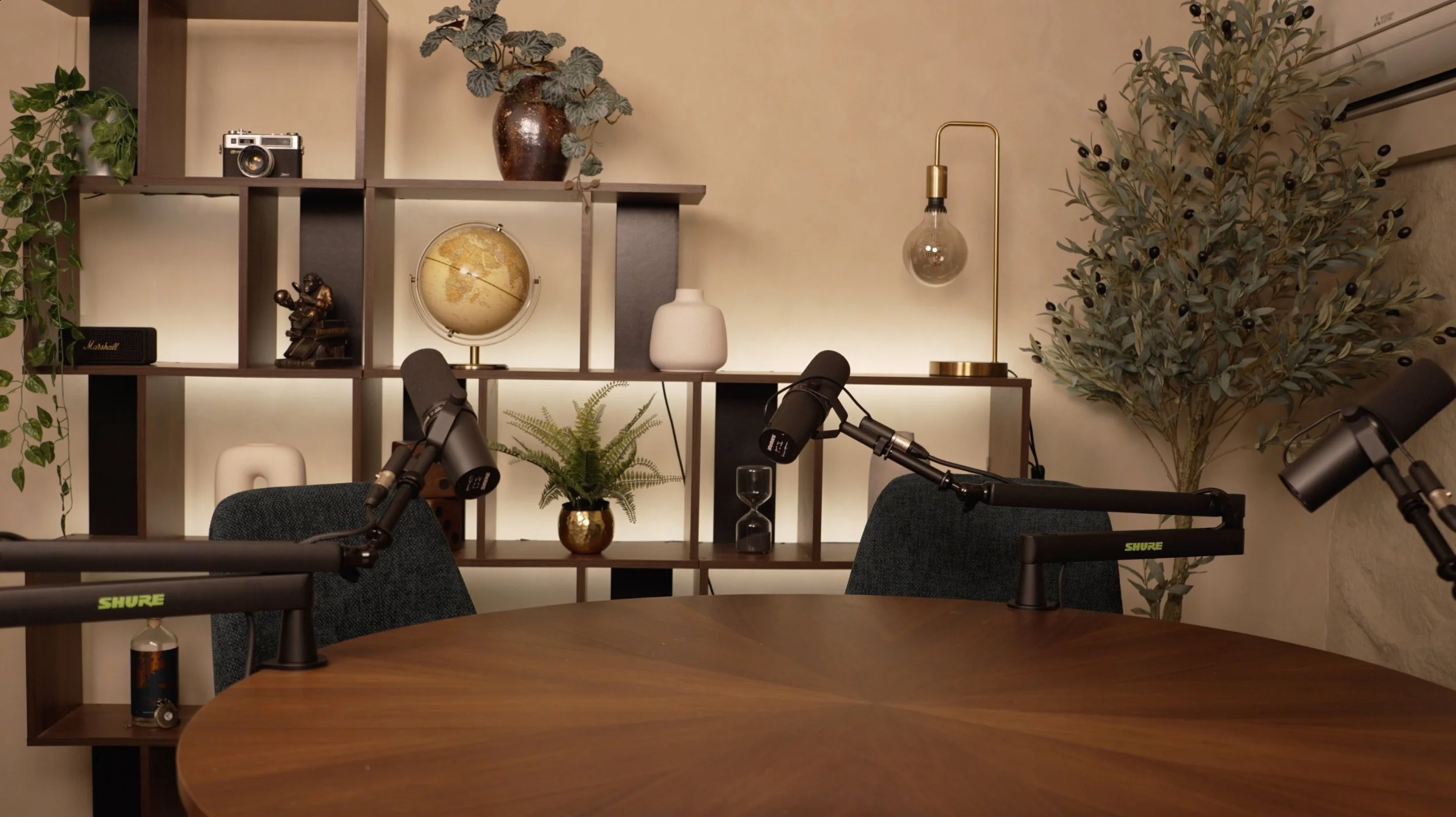 Studio Analogue, a boutique podcast recording Studio in Singapore, with a round table, black microphones, and a background shelf with decorative plants, a globe, a white vase, a statue, and a lamp. 
