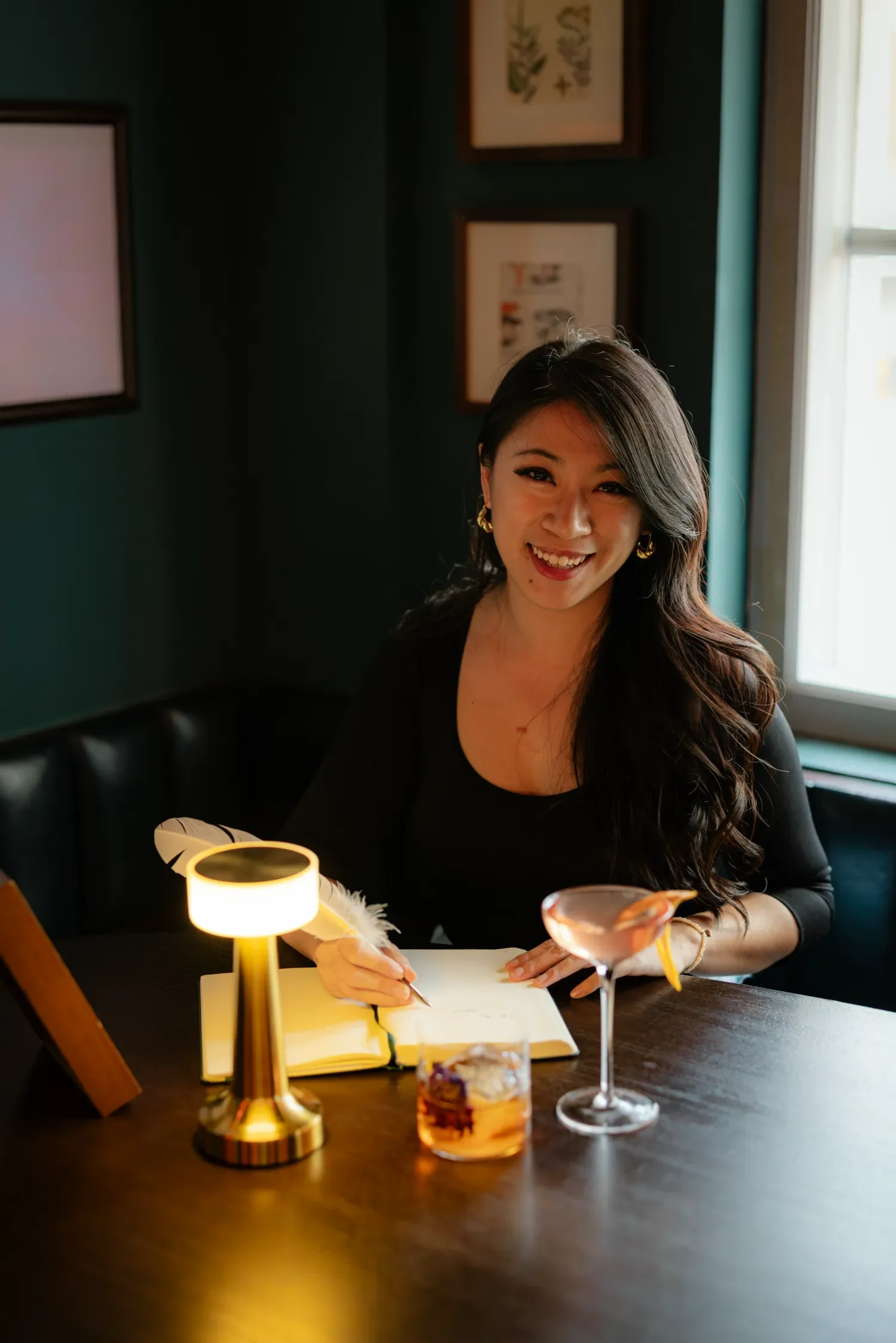 Cheryl Lau, founder of Studio Analogue and award winning podcaster. She is smiling, writing, and wearing gold earrings and a black top. There are framed pictures on the teal-colored wall behind her, and sunlight streaming in through a window.