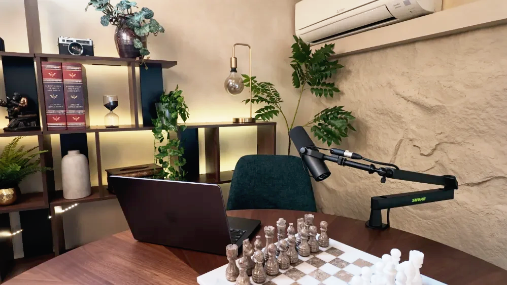 A home office or recording studio with a wooden desk, laptop, chess set, and microphone, with shelves holding books, plants, a vintage camera, and decorative items, and a wall-mounted air conditioner.
