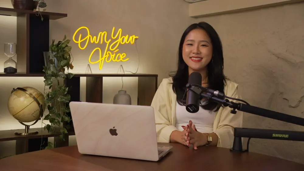 Rae Fung, Speaker, Coach and Professional Host smiling at Studio Analogue, a podcast studio. She have customised the podcast recording studio with a neon sign saying 'Own Your Voice' in the background for her podcast show.