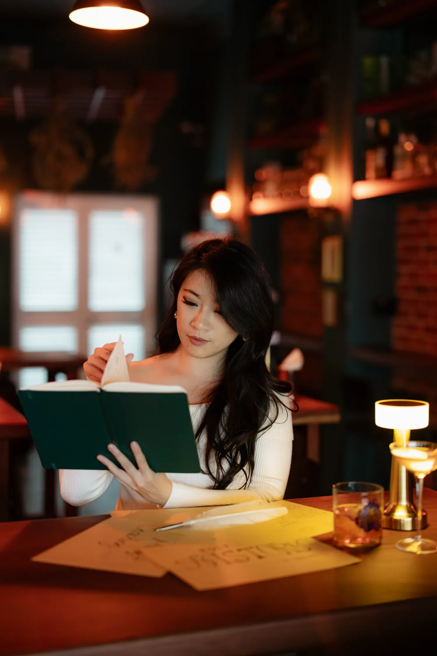 Cheryl Lau, Singapore podcast producer with long dark hair reading a book in a warmly lit restaurant or cafe, with candles and handwritten notes on the table.