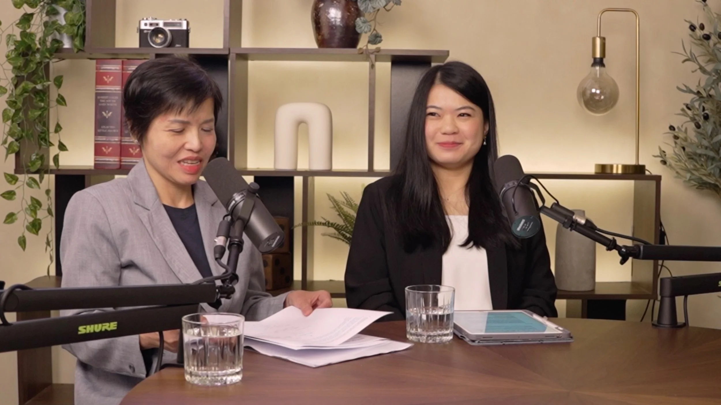 Two women are sitting at a table with microphones, glasses of water, papers, and a tablet, recording a podcast or radio show in a cozy decorated room.