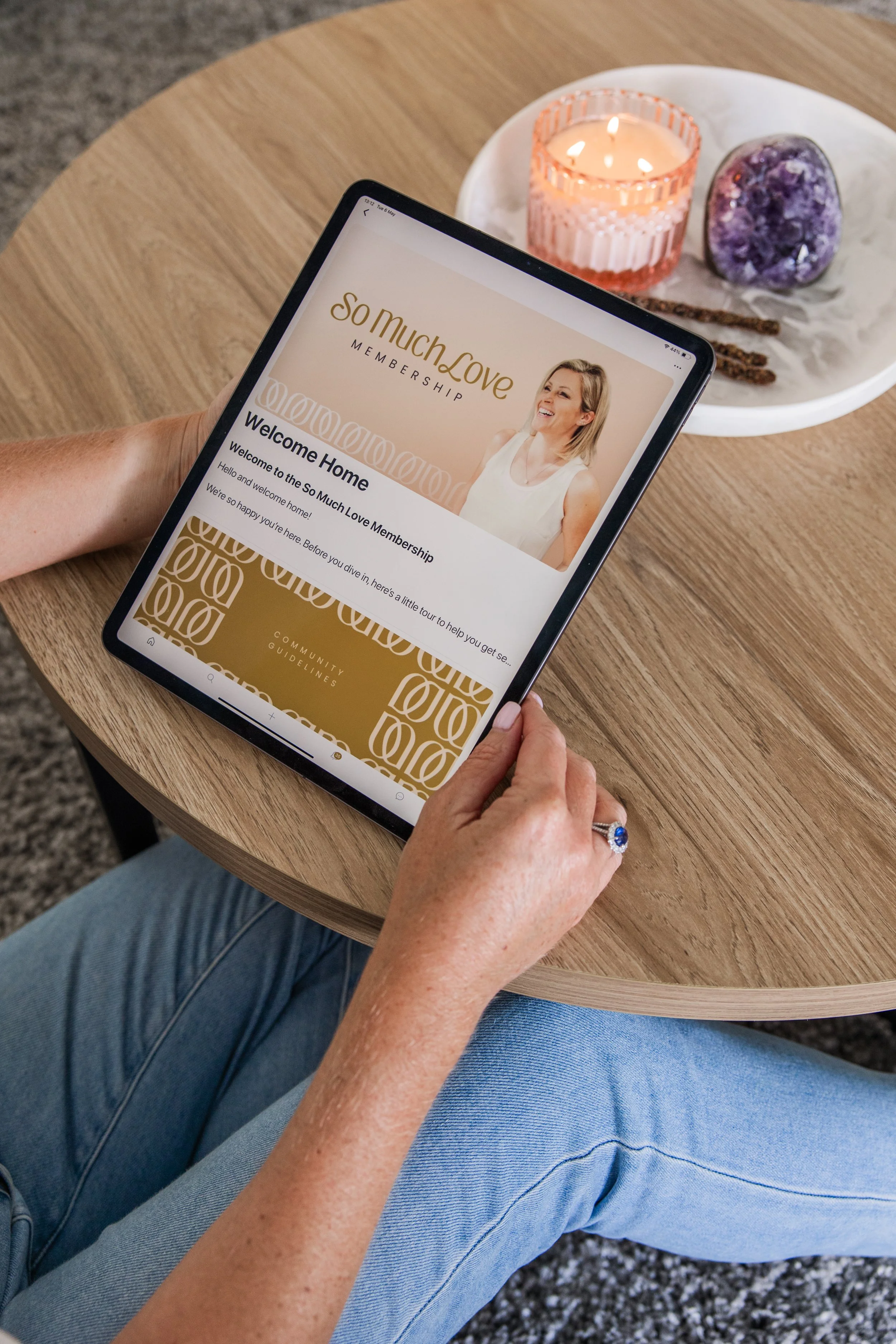 Person sitting at a wooden table holding a tablet displaying a welcome message for a membership called So Much Love. On the table, there is a white tray with a lit pink candle, a purple amethyst stone, and two sticks. The person is wearing blue jeans and has a ring on their finger.