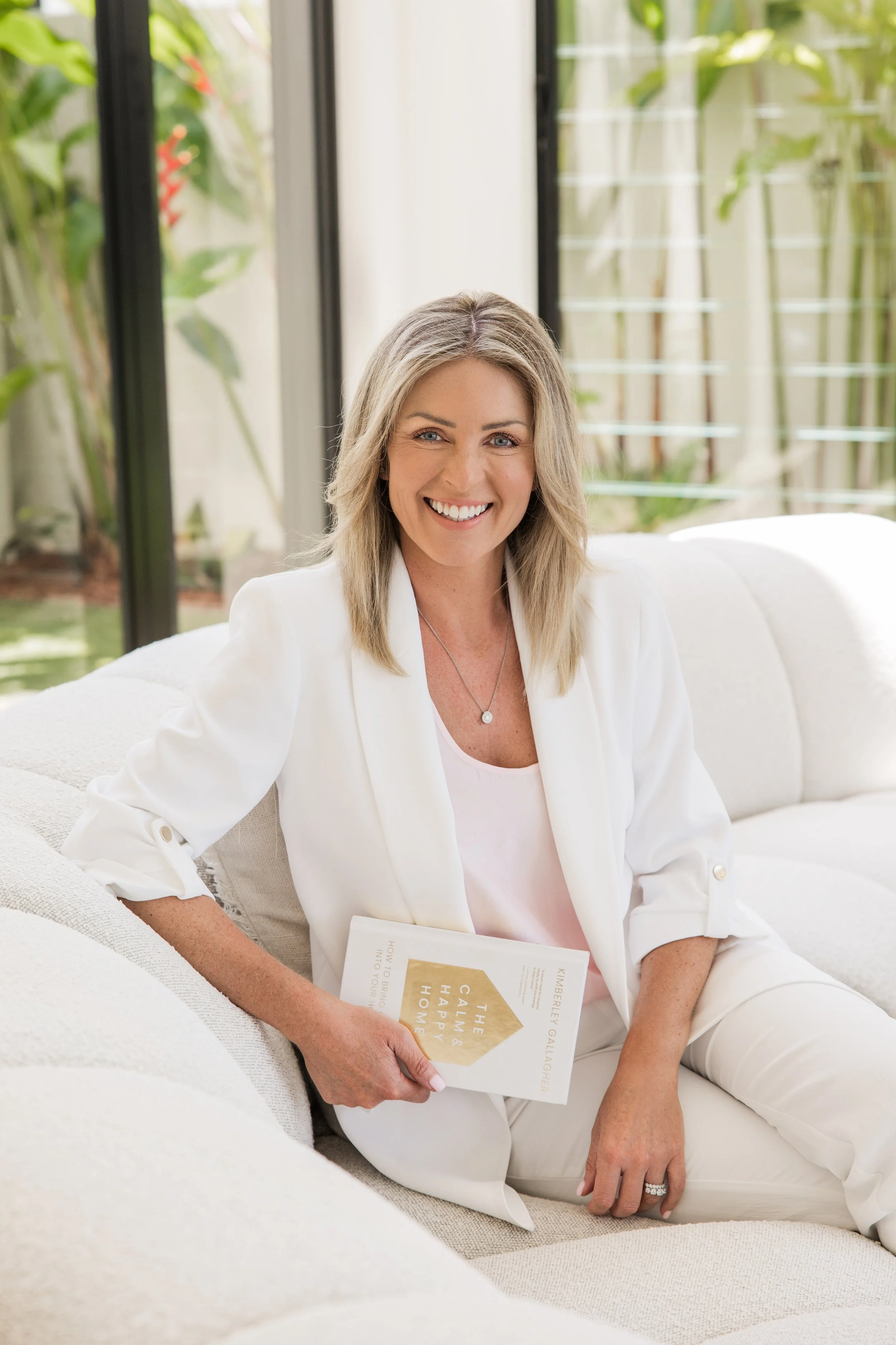 Kimberly Gallagher wearing a white blazer and pants, sitting on a white couch holding a book, smiling in a bright room with large windows and greenery outside.