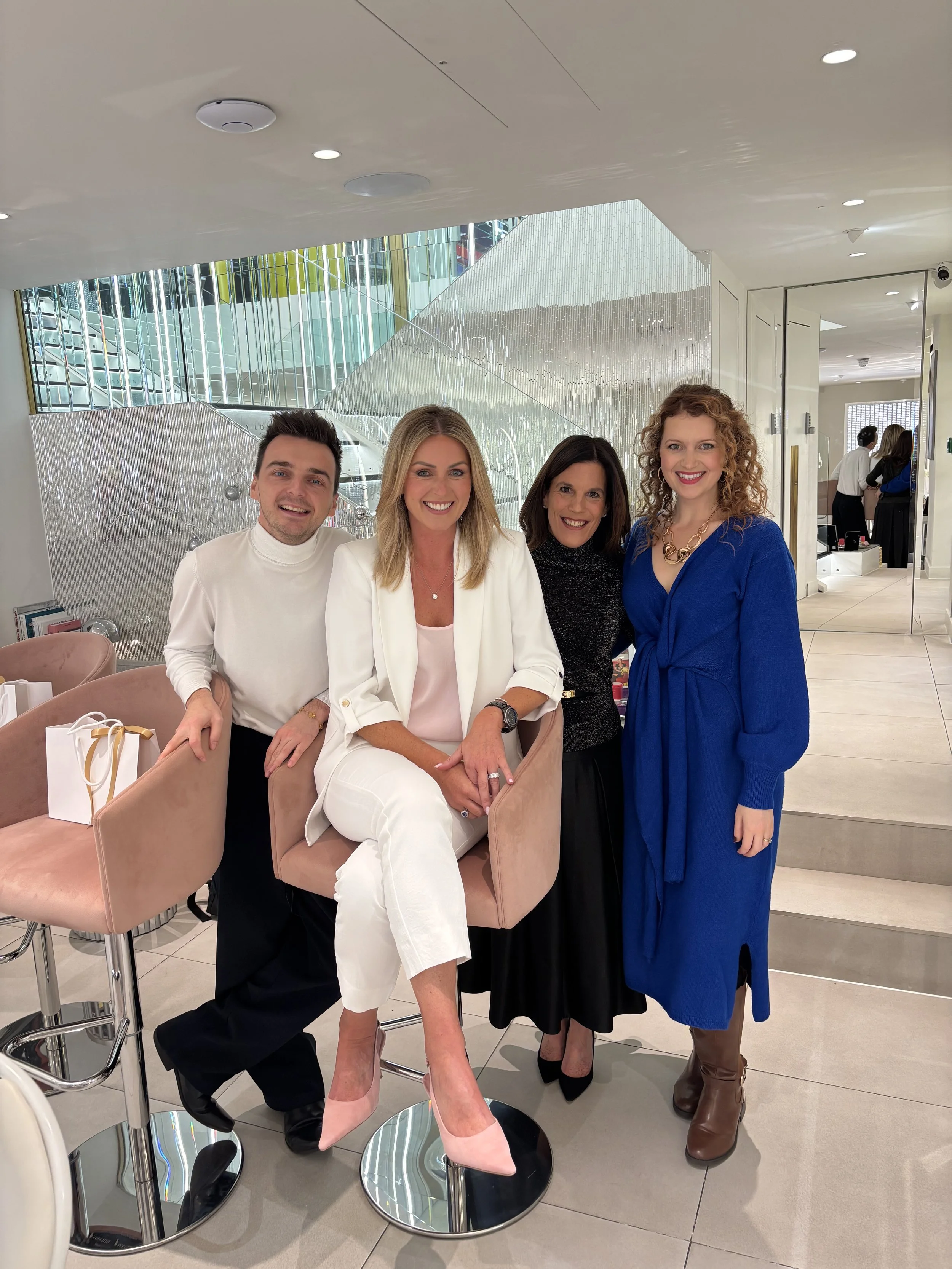 Four smiling people, three women and one man, posing together indoors in a modern, well-lit space with a reflective staircase in the background. The woman in the white blazer is seated on a high stool, while the others stand around her.