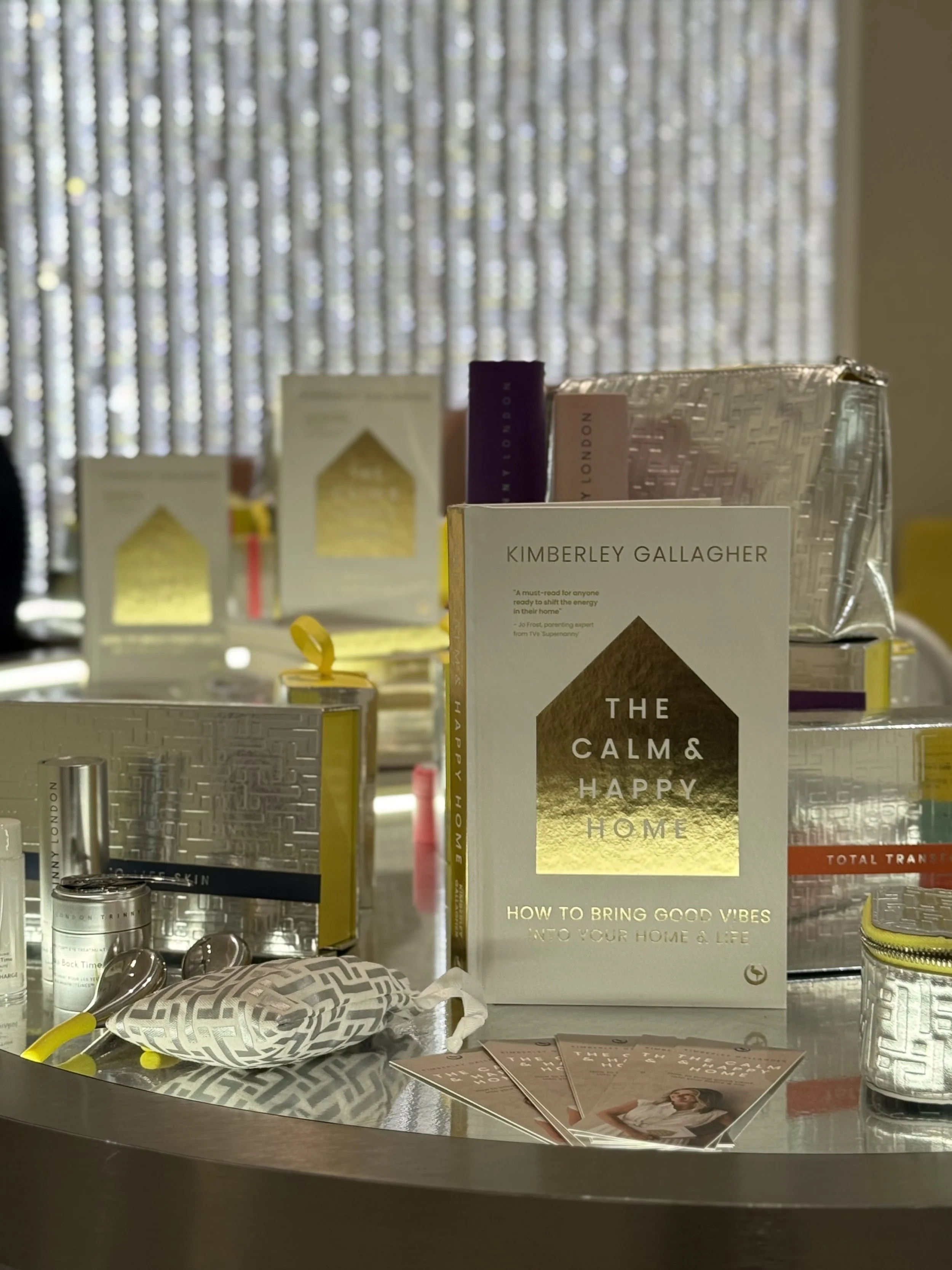 Display of various items on a table, including books, skincare products, brochures, and decorative objects, set against a backdrop of blue curtains with white dots.