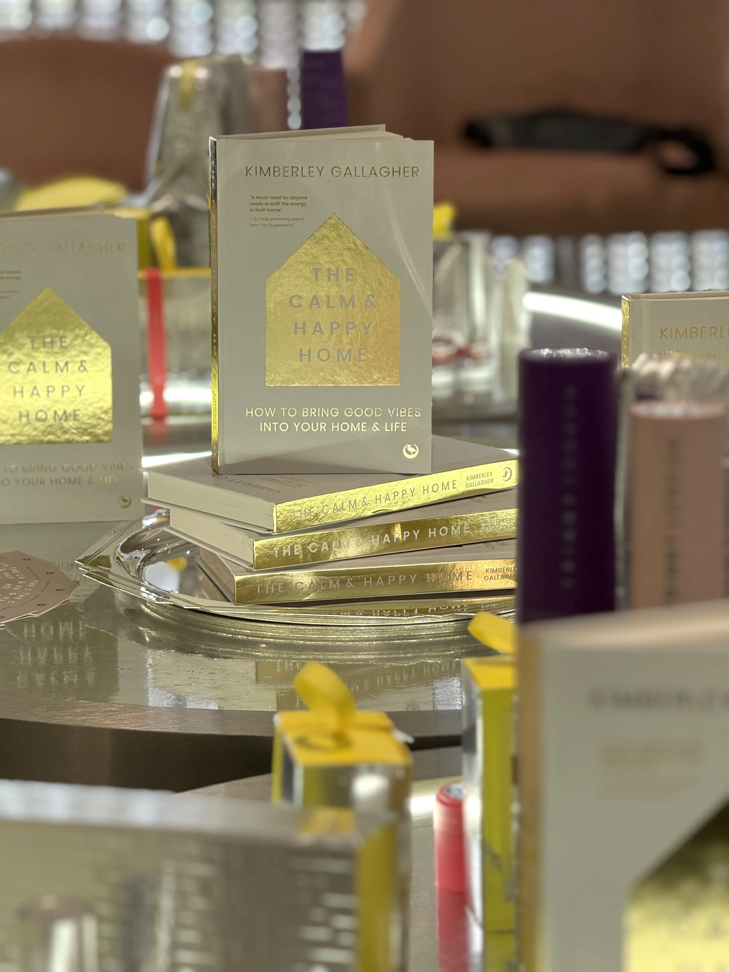 Stack of books titled 'The Calm & Happy Home' by Kimberley Gallagher displayed on a silver tray, with colorful small candles and blurred background.