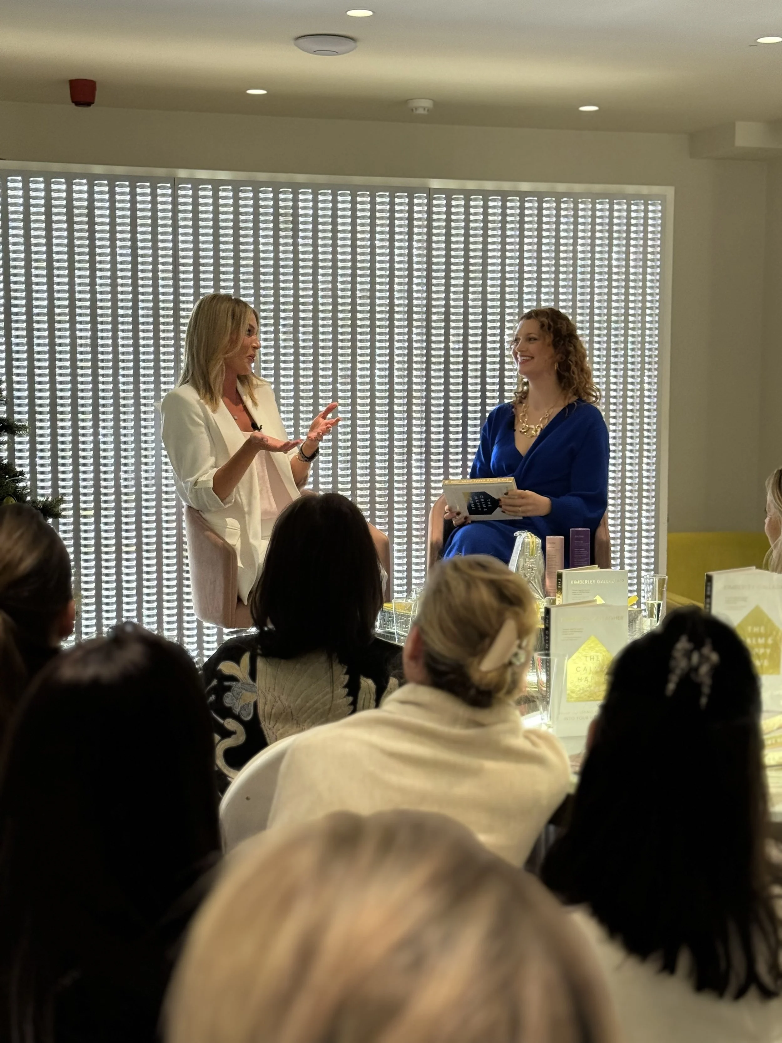 Two women are engaging in a conversation or interview, one standing and speaking with hand gestures, the other sitting and smiling, holding a book or notepad, in a well-lit room with an audience watching.