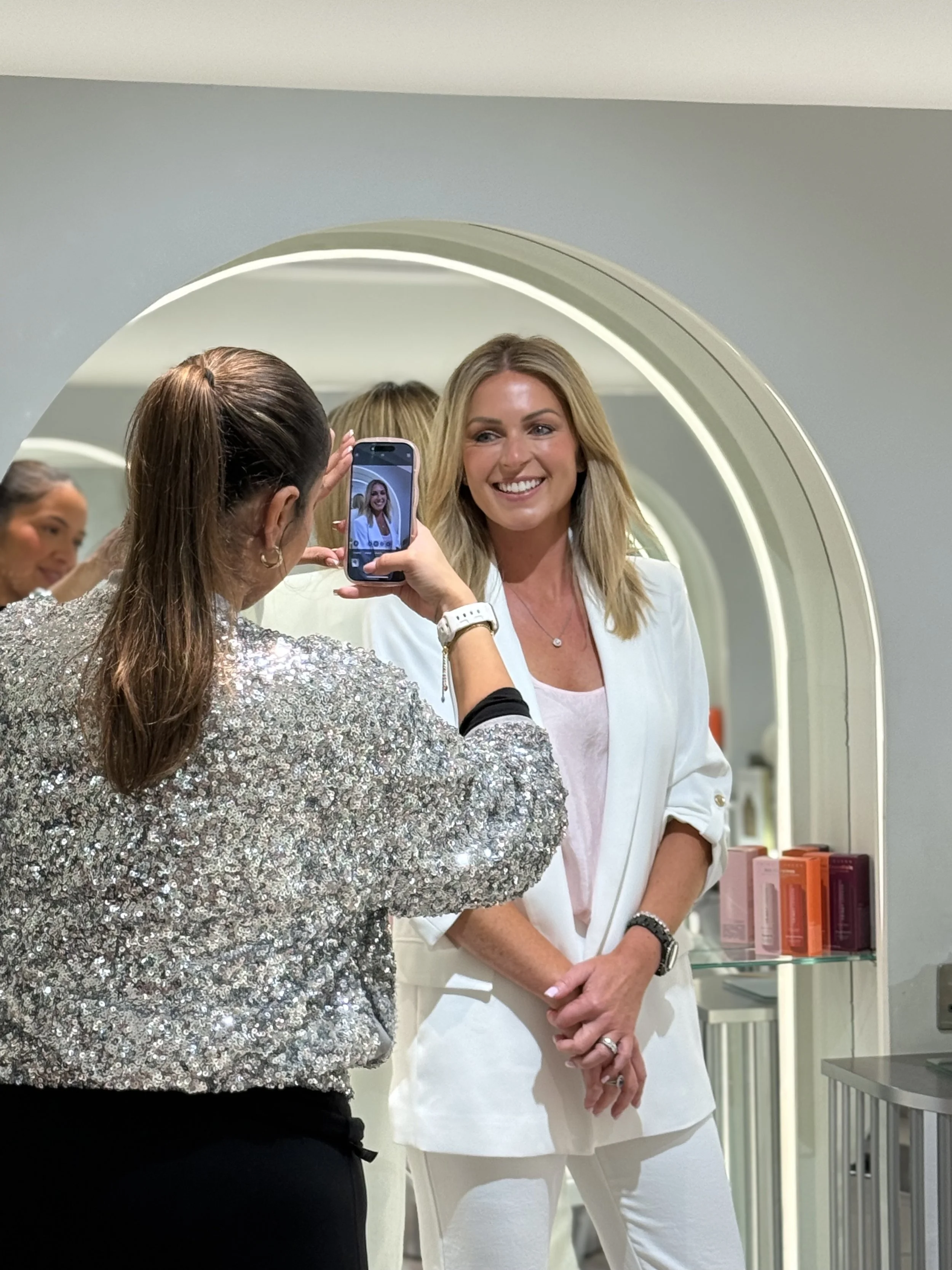A woman with blonde hair dressed in white is having her picture taken in a mirror by another woman in a sparkly top. They are in a modern room with a display of pink and orange skincare or cosmetic products.