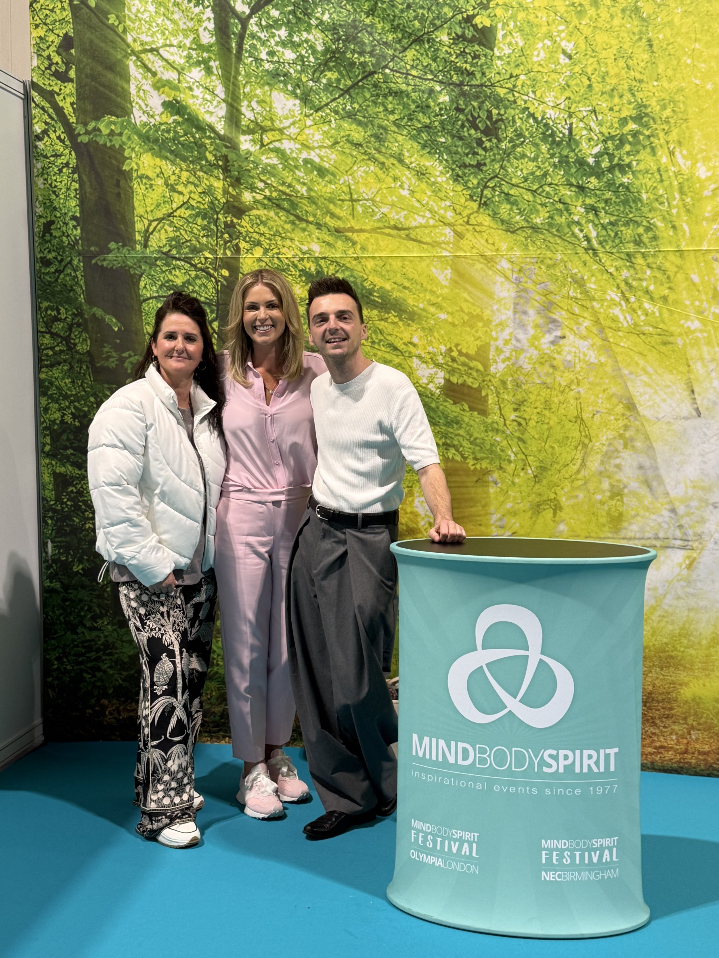 Three people standing together at the Mind Body Spirit Festival, with a backdrop of a green forest and sunlight. Two women and one man are smiling, with a turquoise display table in front of them bearing the festival's logo and locations.