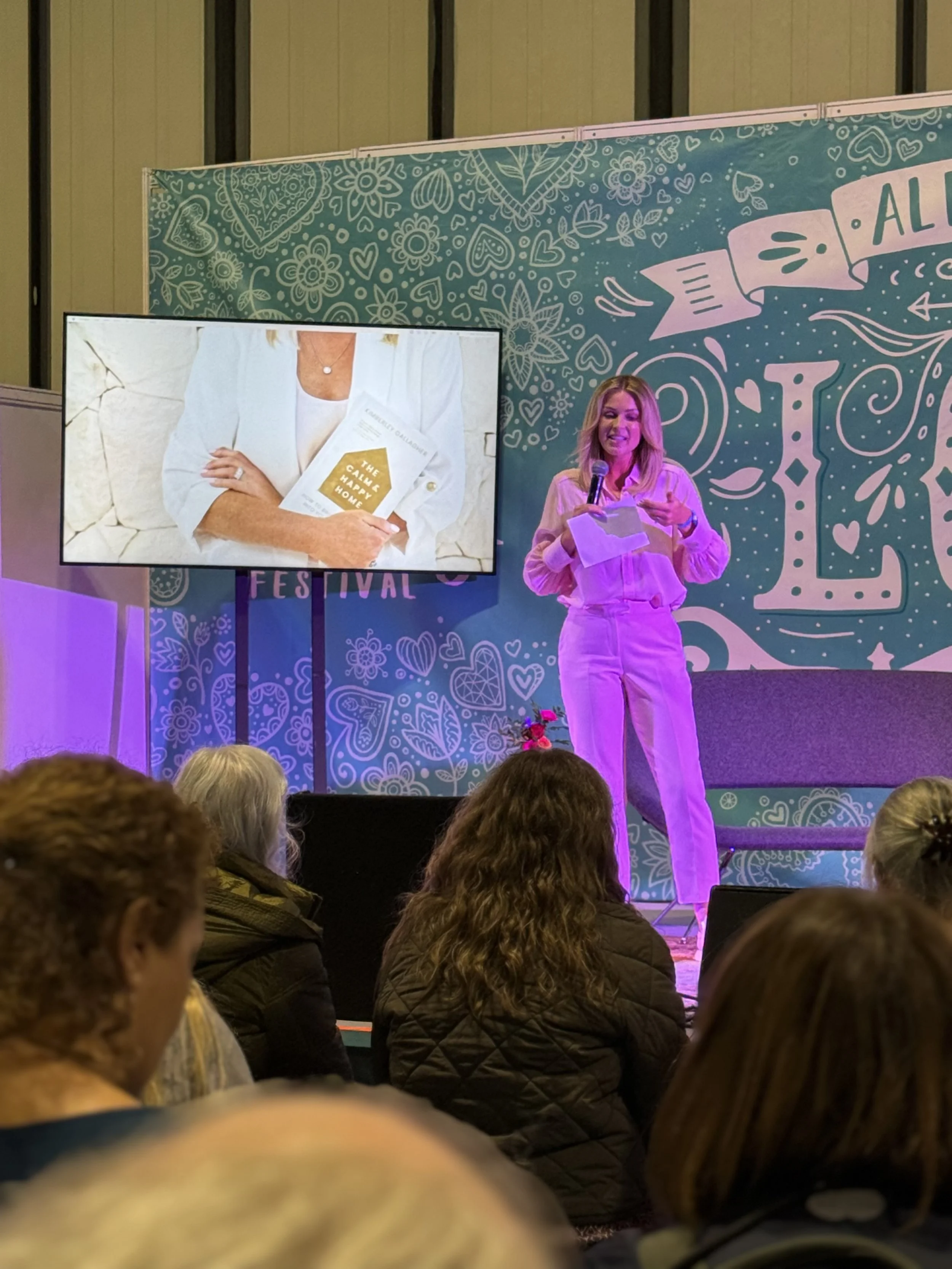 A woman with blonde hair, wearing a white outfit, is standing on stage with a microphone, reading from a piece of paper. Behind her, there is a large decorative blue backdrop with white patterns and the word 'LOVE.' A screen on the left shows a close-up of a person holding a booklet and wearing a white blazer. Audience members are visible in the foreground.