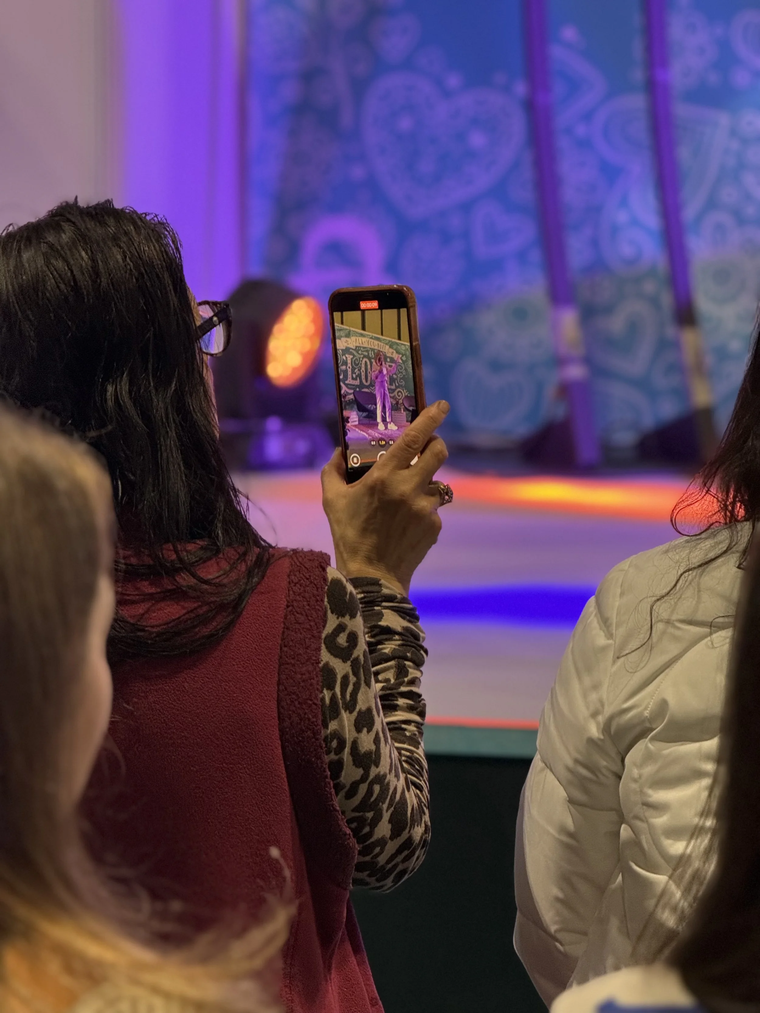 Person holding a smartphone recording or taking a picture of a performer on stage during an event or show. Audience members are visible in the foreground, with a colorful, patterned backdrop behind the stage.