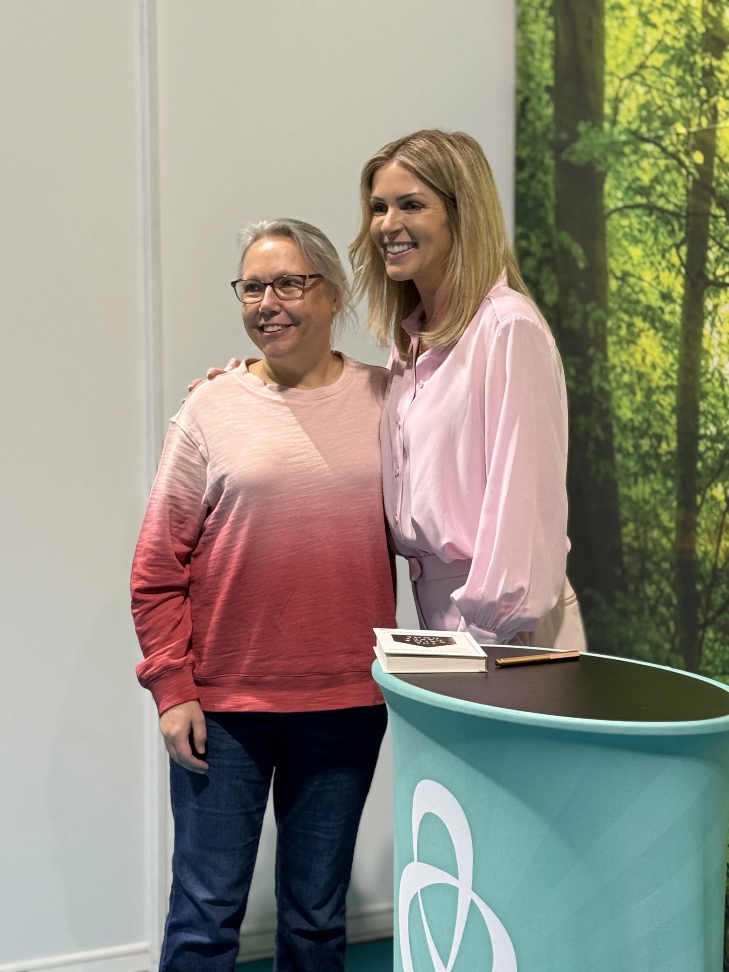 Two women standing together, smiling, near a teal-colored table with a book and a pen on top, in front of a background with a nature scene of trees and greenery.