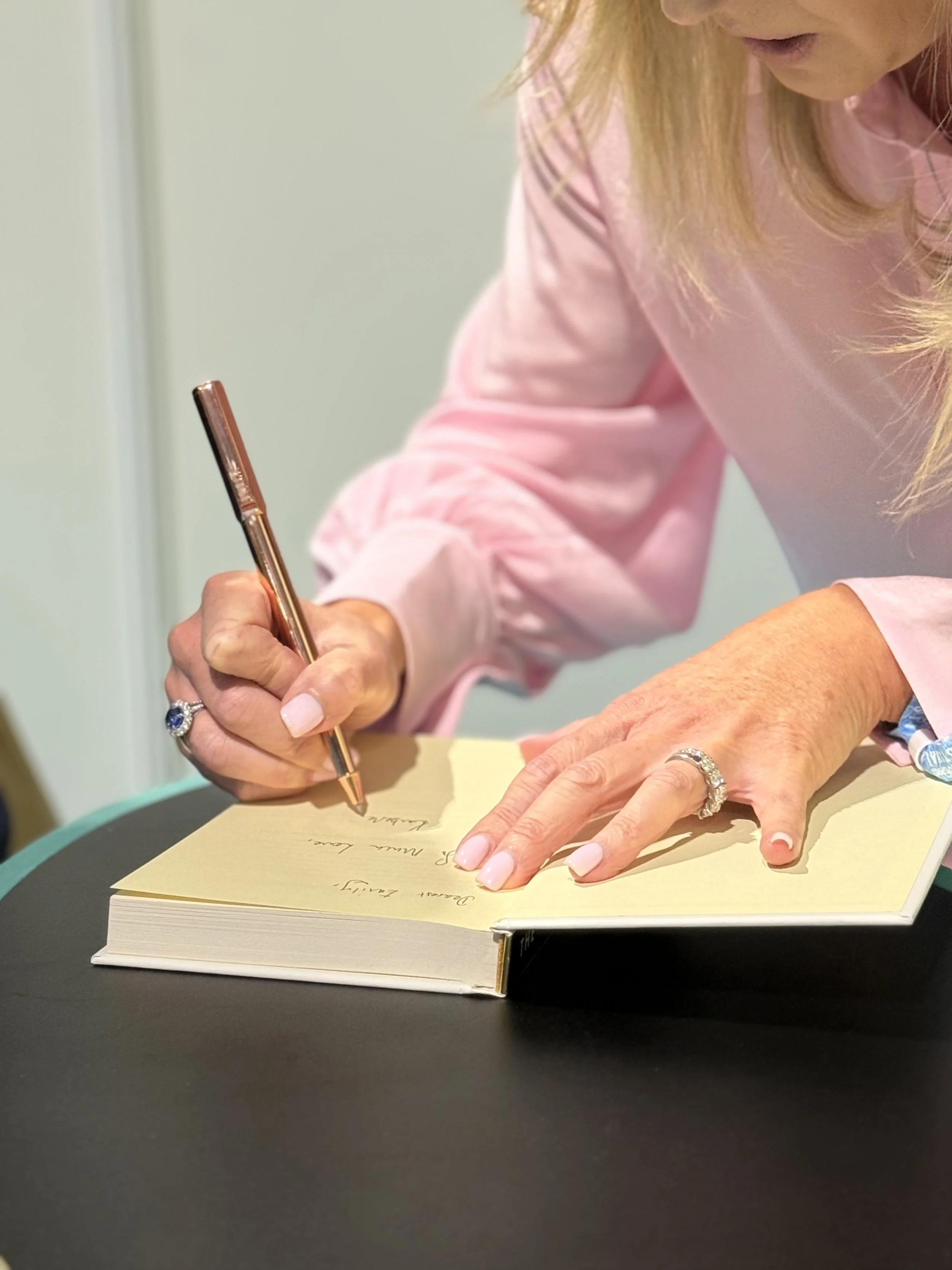 A woman in a pink shirt is signing a book with a gold pen. She is wearing rings with blue stones and has long blonde hair. The book is open on a black table.
