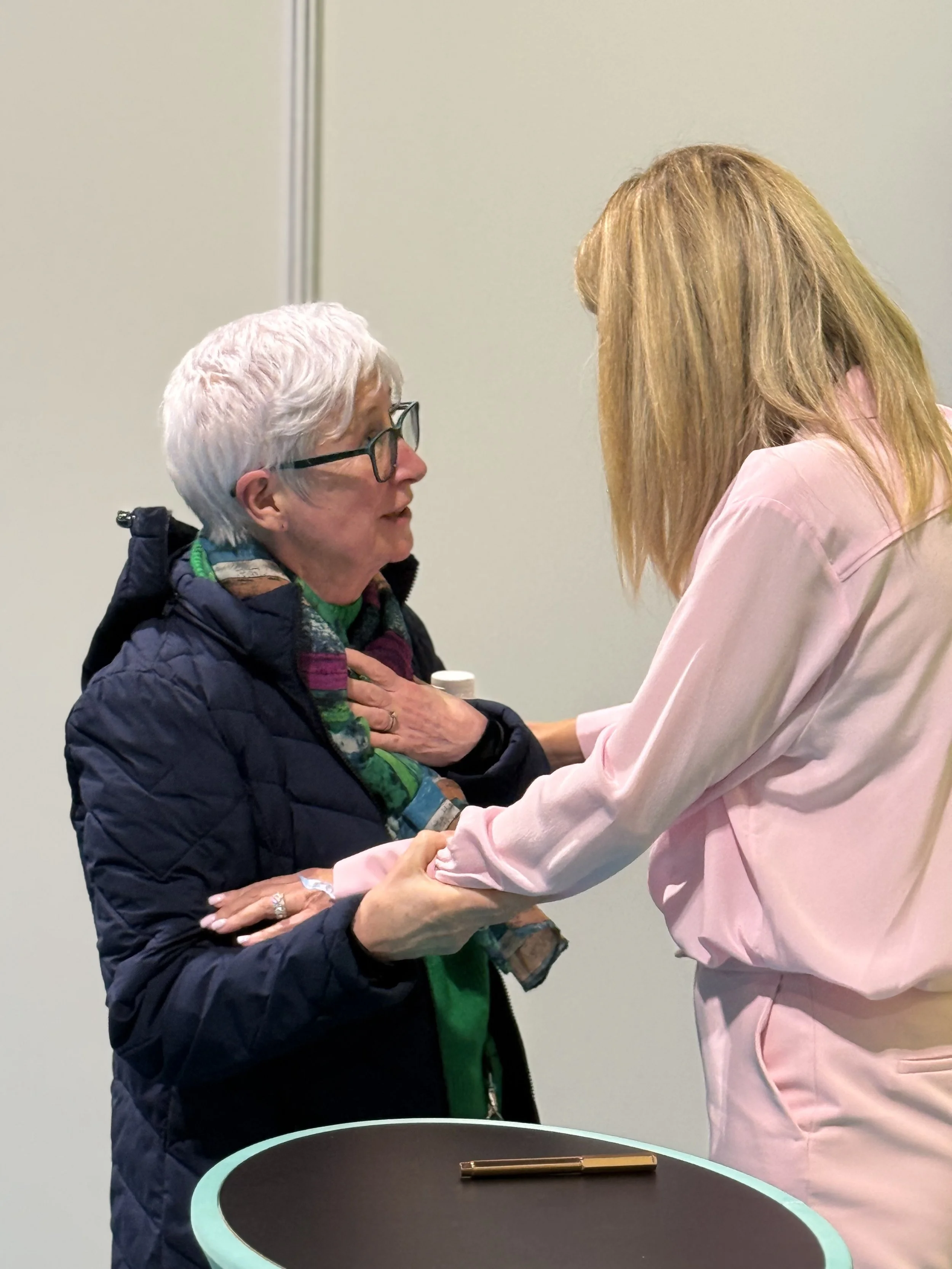 An elderly woman with white hair, glasses, and a dark jacket, engaging in a close, personal conversation with a woman with long, reddish-blonde hair wearing a light pink jacket. They are standing near a round table with a black surface and a gold pen resting on it, against a plain beige wall.
