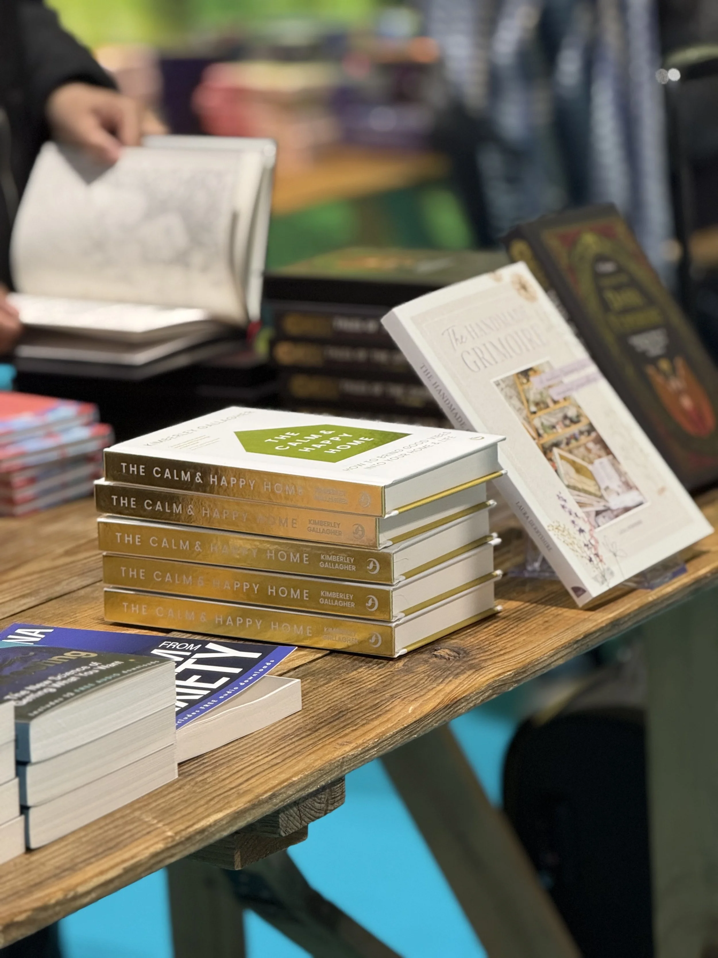 A wooden table displaying several stacks of books, including titles like 'The Calm & Happy Home' and 'The Hanuman Temple'. In the background, blurred people are browsing more books at what appears to be a book fair or bookstore.