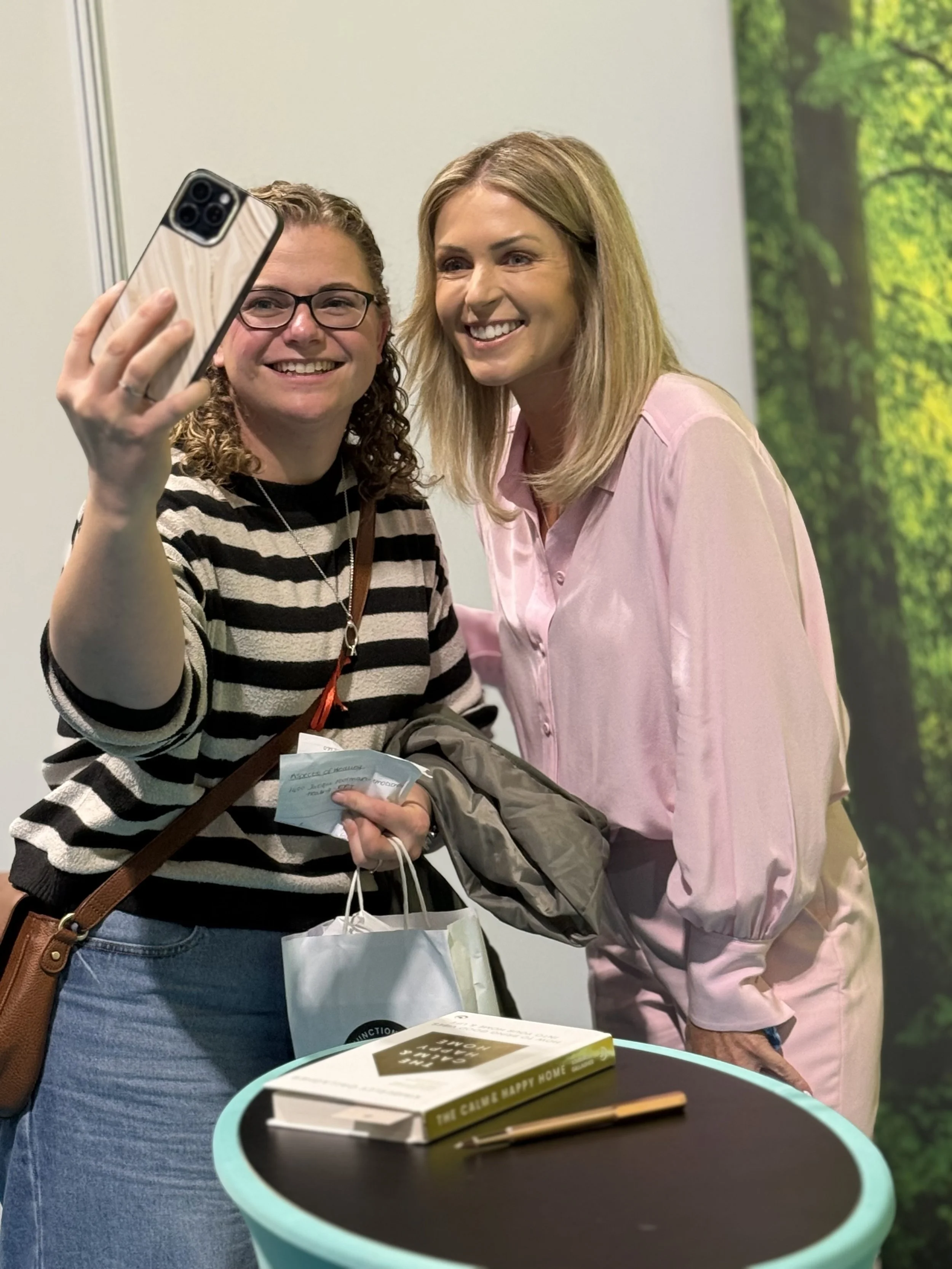 Two women taking a selfie together indoors, one wearing glasses and a striped shirt, the other with blonde hair and a light pink shirt.