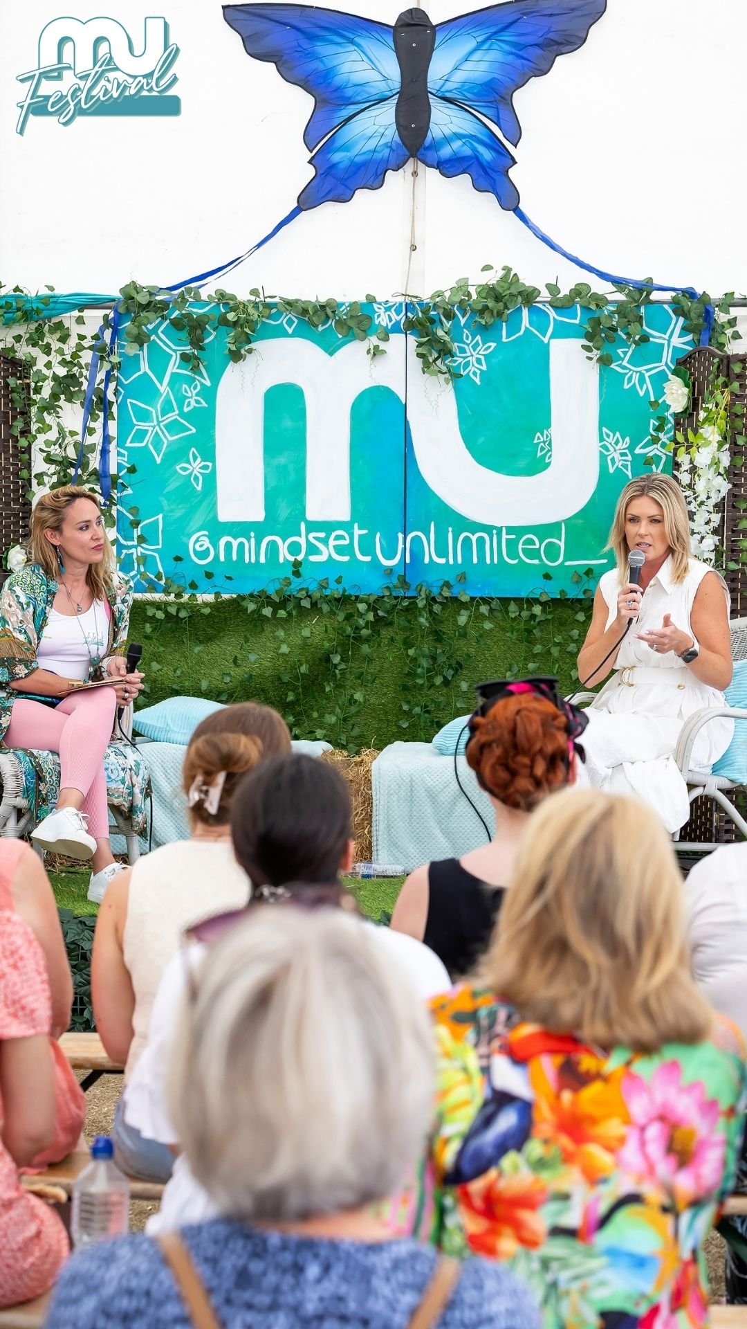 A woman in a white dress speaking into a microphone at an outdoor event with an audience of diverse women wearing colorful clothing. Behind her is a large blue butterfly decoration and a sign with the text 'miU' and '@mindsetunlimited', surrounded by green plants and flowers.