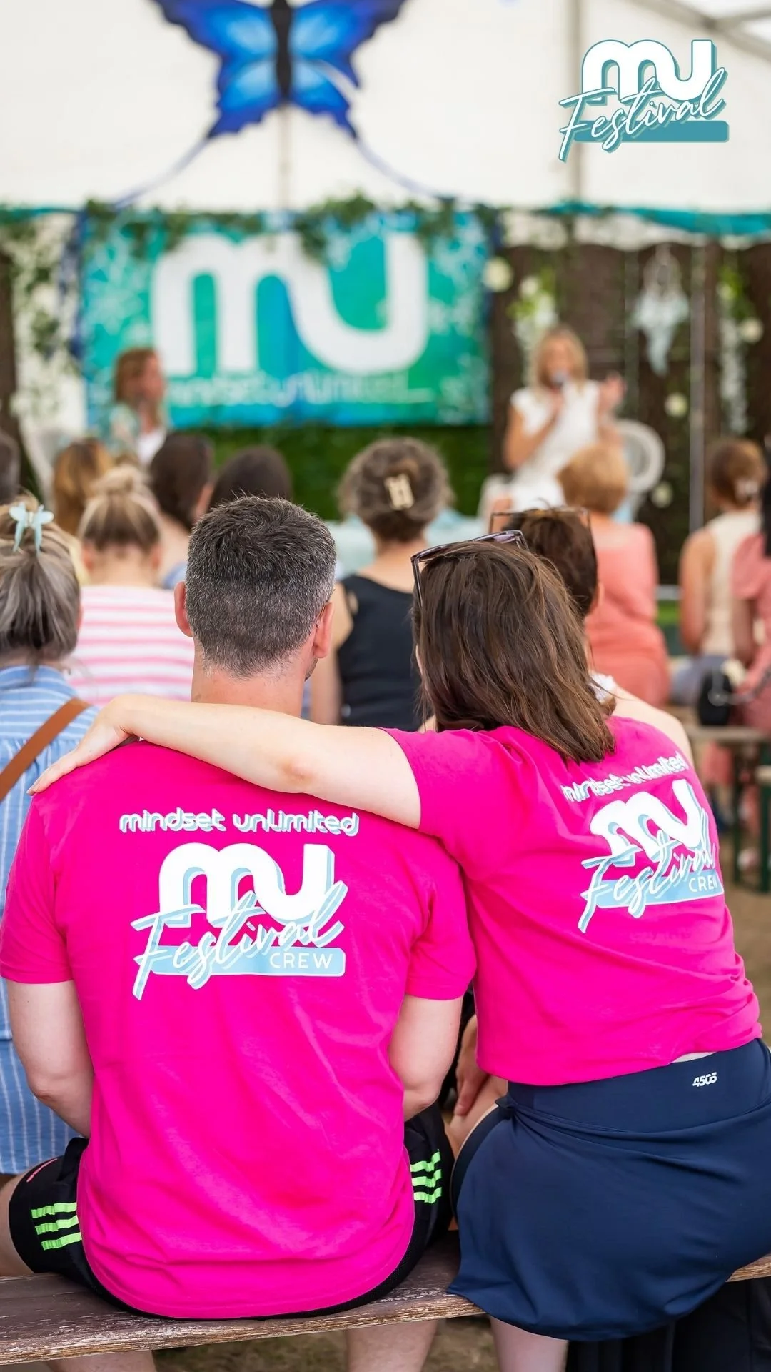 Two people wearing bright pink t-shirts with 'mindset unlimited MCX Festival Crew' sitting close together, with one arm around the other's shoulders, watching a presentation at an outdoor festival.