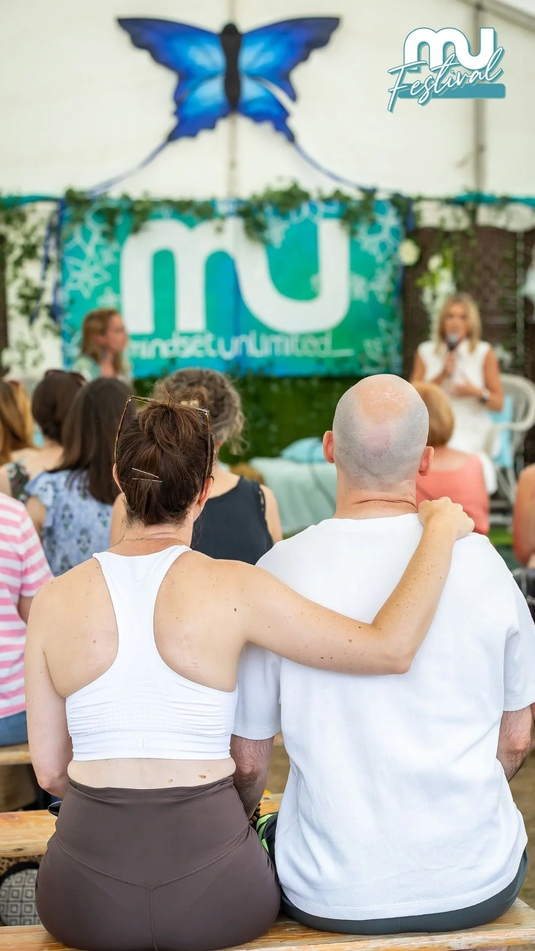 People attending a live outdoor event with a Ukrainian theme, sitting on benches, with a large projection screen in the background and a woman giving a speech.