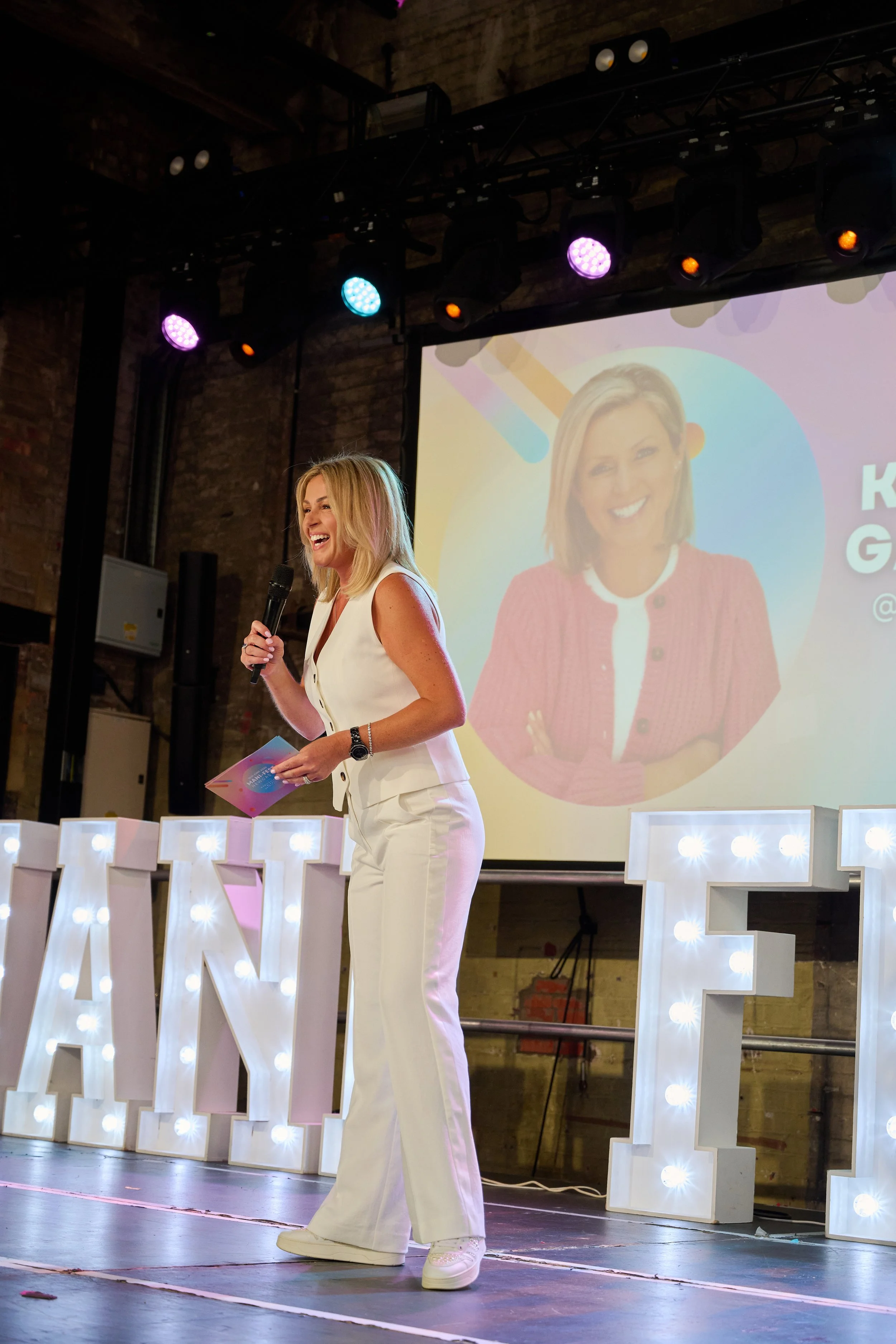 Kimberly Gallagher standing on stage holding a microphone and a  booklet, smiling. Behind her, there are large illuminated letters spelling out part of a word, and a large screen displays a picture of her with a rainbow background and some text.
