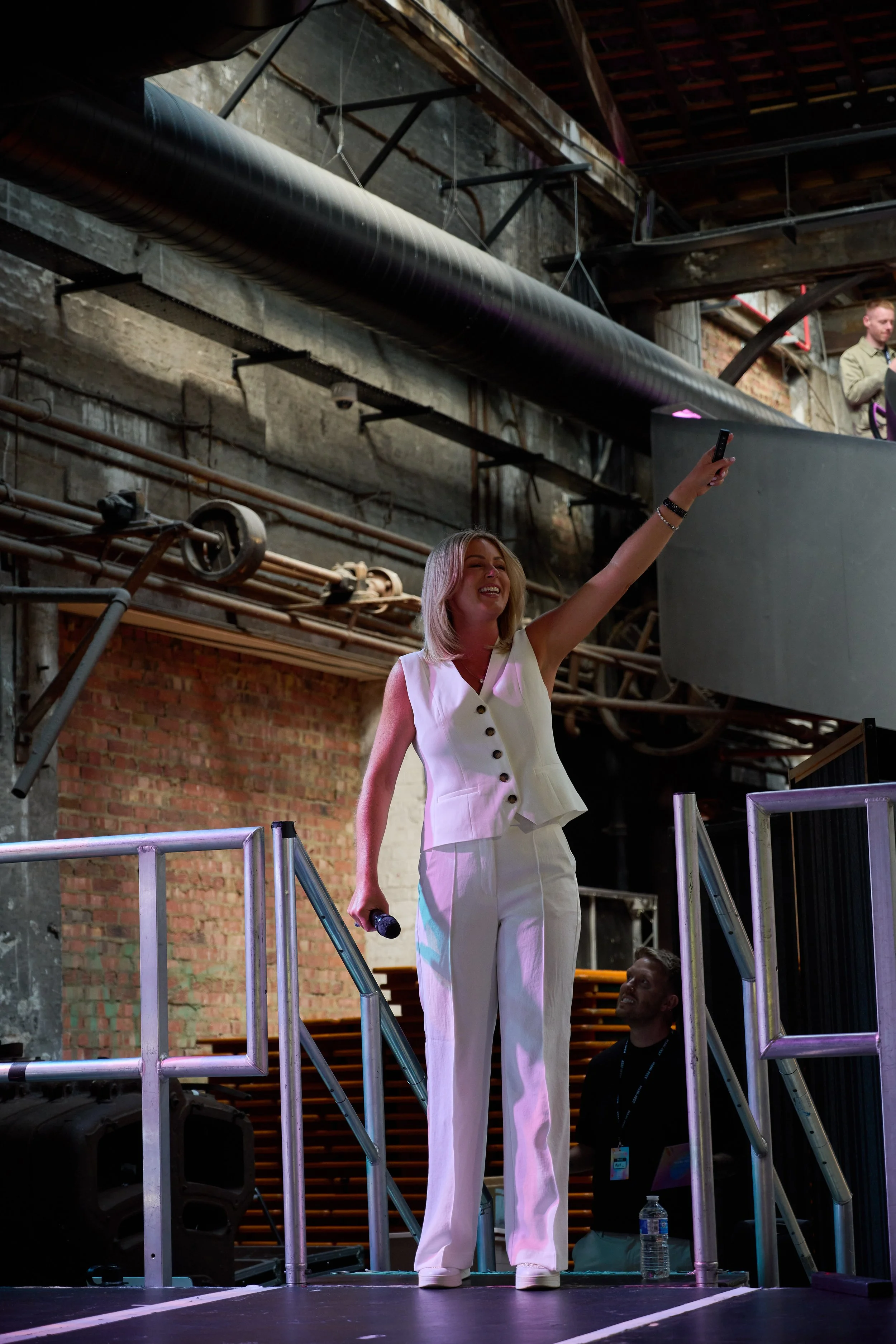 A woman in a white sleeveless outfit is standing on an industrial-style stage, holding a microphone in her right hand while smiling and waving with her left hand. She is looking at the crowd. A man is sitting below the stage, looking up at her, with a water bottle on the floor nearby. The background has exposed brick walls, pipes, and metal structures.