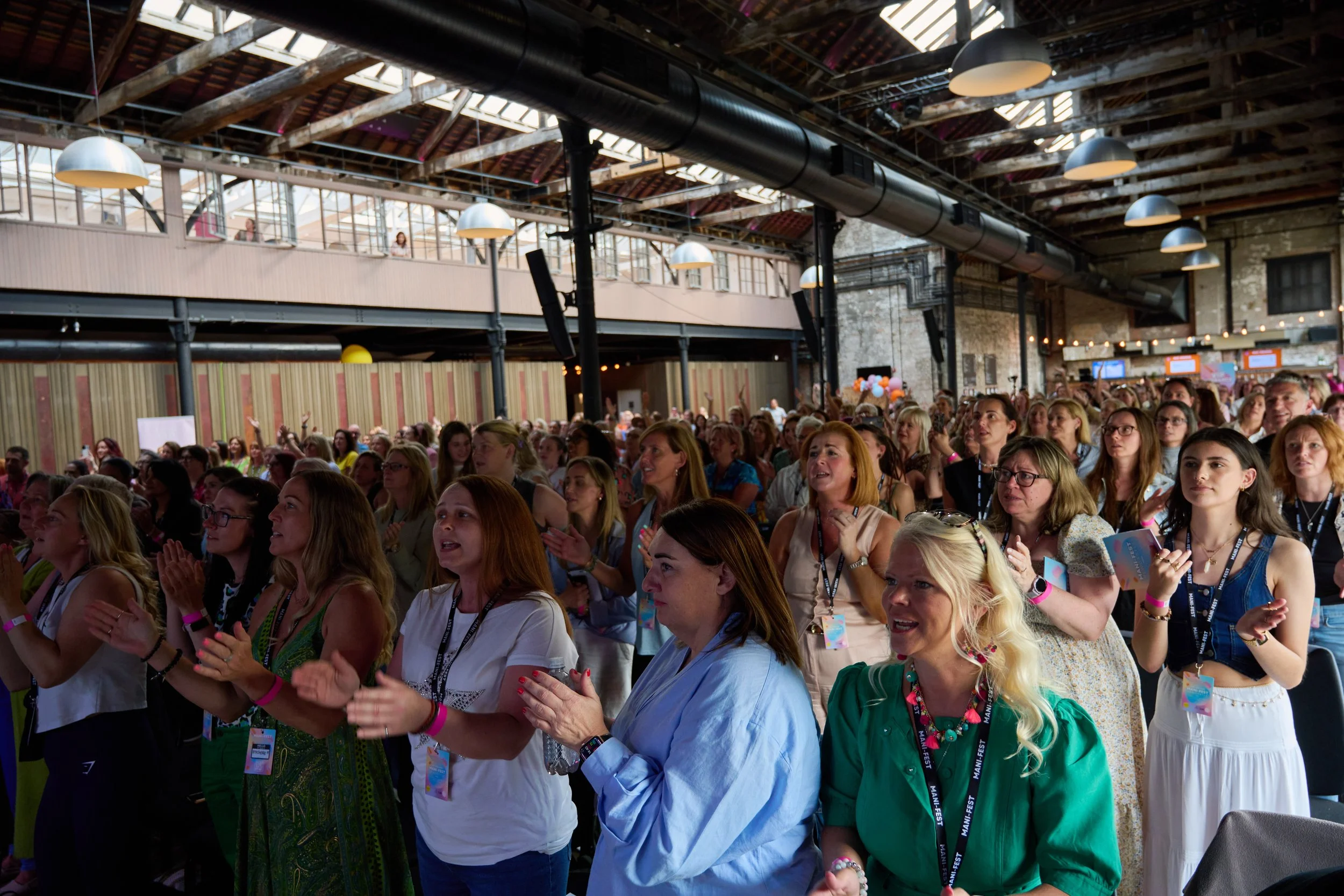 A large crowd of women attending an indoor event, clapping and looking towards the stage. The venue has a high, exposed ceiling with wooden beams, skylights, and industrial lighting fixtures. Some women are wearing badges and pink wristbands.