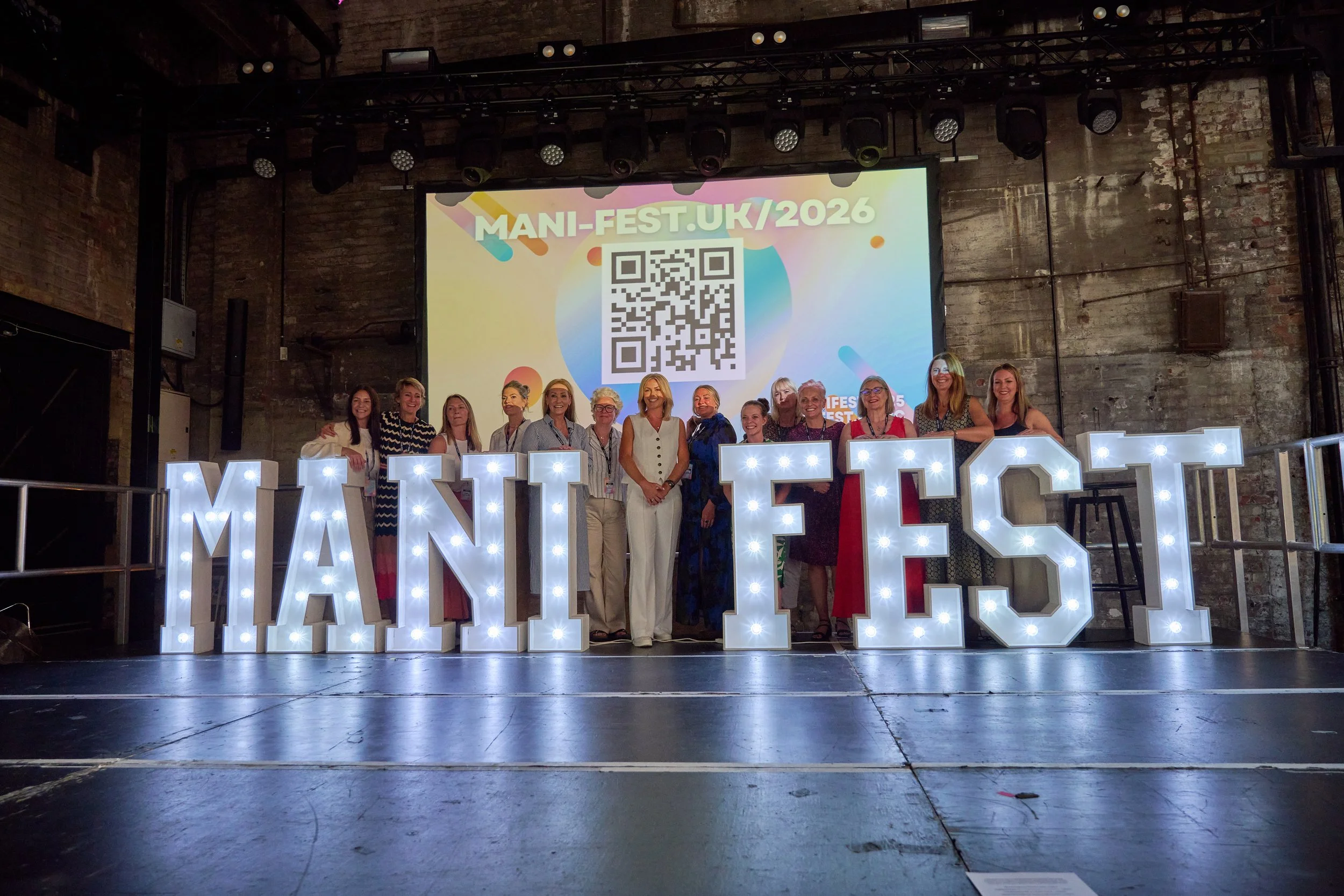 Group of women standing together on stage at Manifiest UK 2026 event, behind large illuminated letters spelling 'MANIFEST' and a large screen displaying event information with a QR code, in an industrial-style venue with brick walls and stage lighting.